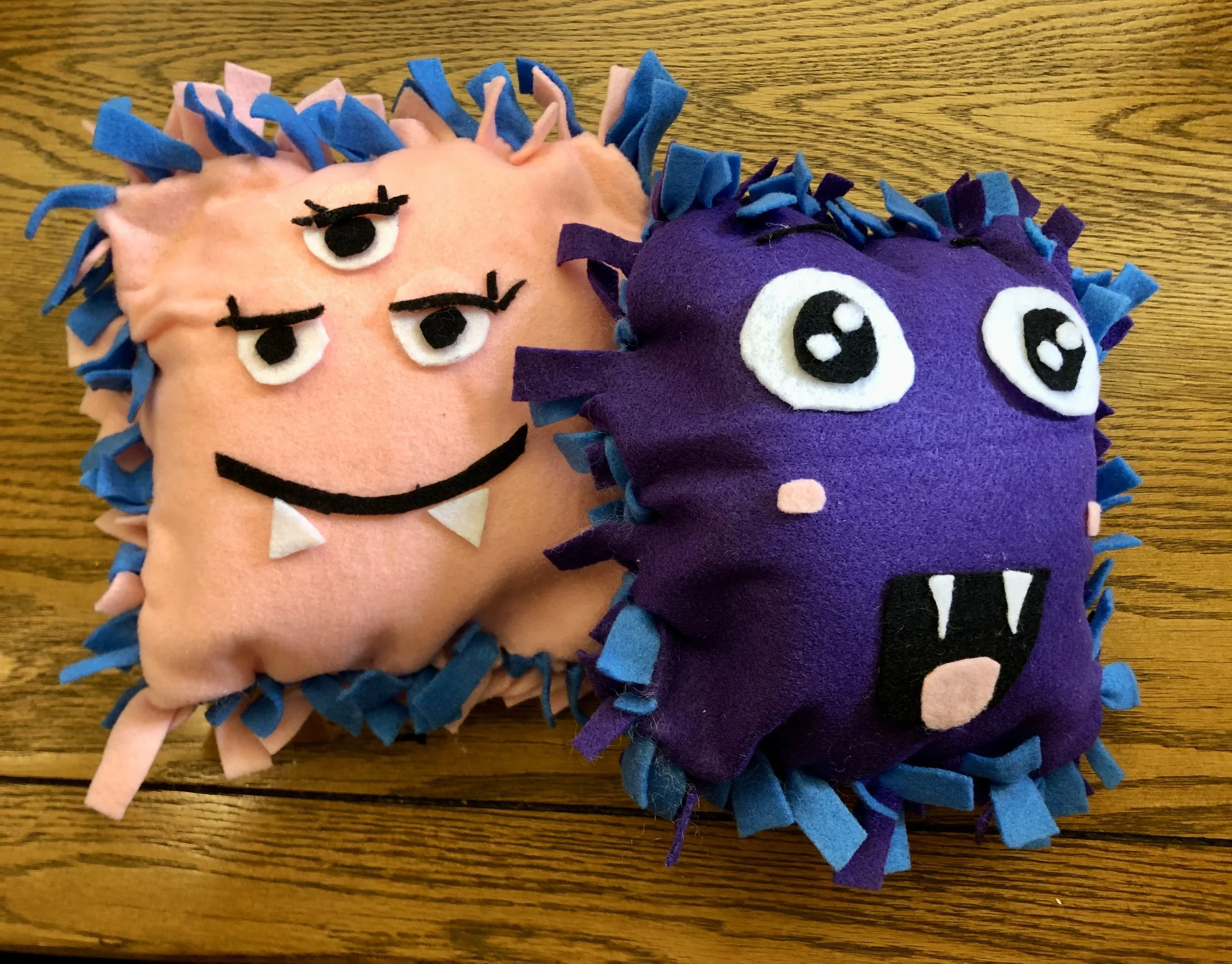 Two handmade plush toys resembling cartoon faces with spiky hair made of felt, one pink with blue hair and one purple with blue hair, placed on a wooden surface.