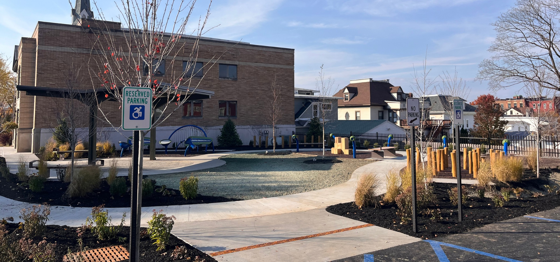 Accessible children's playground with sensory play equipment, landscaped paths, and reserved accessible parking signs outside a brick building.
