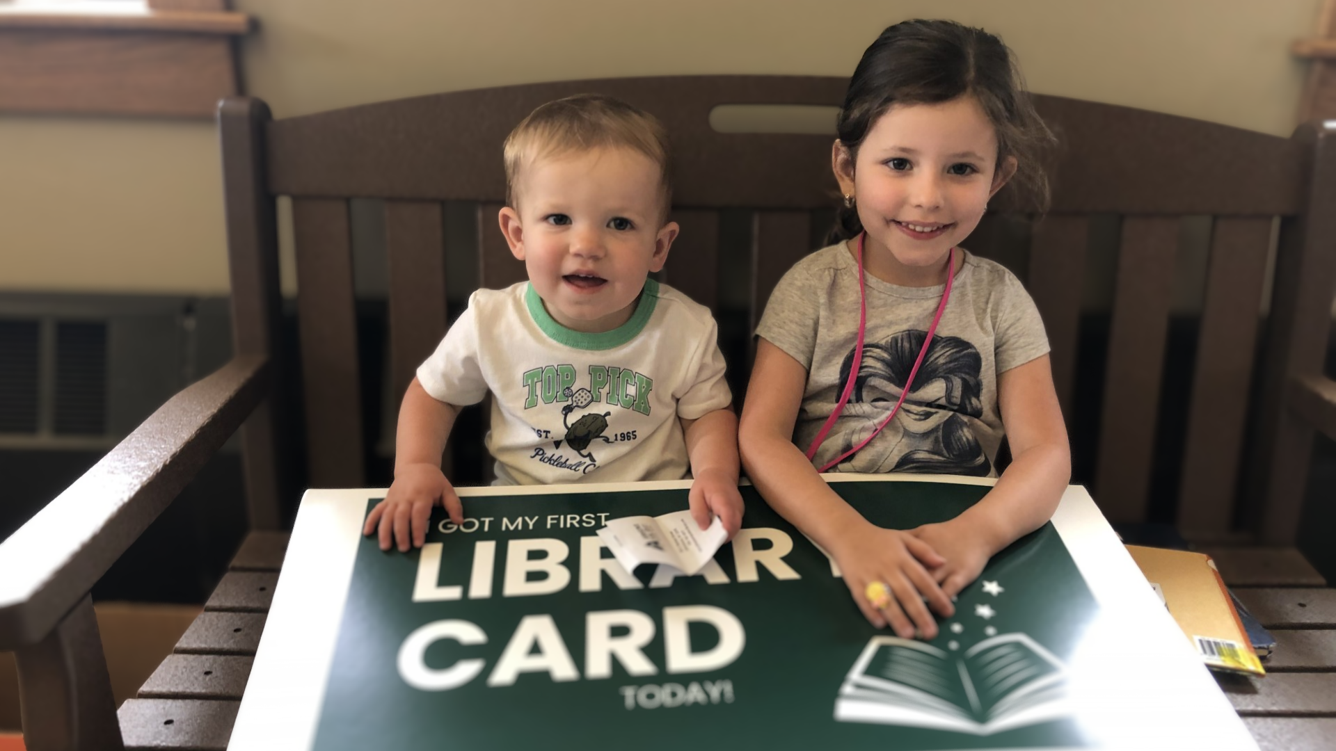 Two children sitting at a table with a sign that reads 'Got my first library card today.' The younger boy has short blond hair, is wearing a white T-shirt with green trim and graphics, and is holding a library card. The girl has curly dark hair, is w