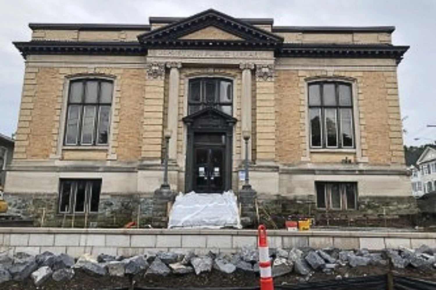 A large, historic brick building with stone details, black door, and big windows, under construction with rocks and safety cones in front.