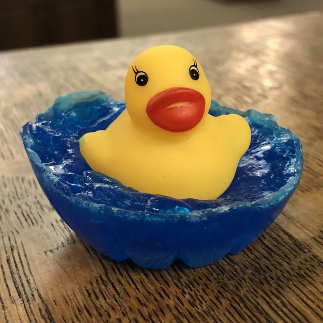 A yellow rubber duck with a red beak and painted eyes floating in a small blue glass dish resembling water, on a wooden surface.