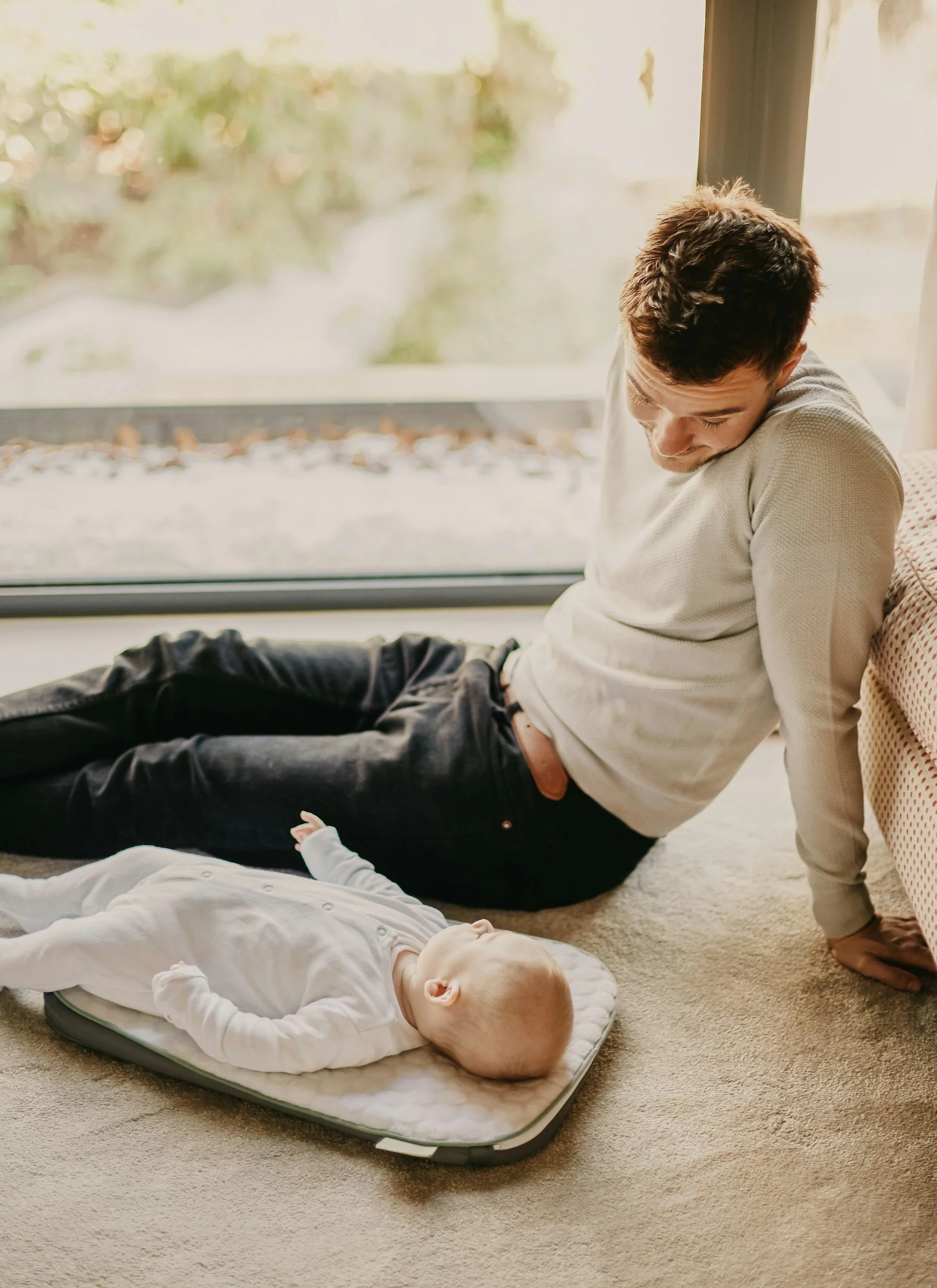 A young man sitting on the floor next to a baby lying on a mat, with a large window in the background showing a blurred outdoor scene. The young man is leaning forward and looking down at the baby.