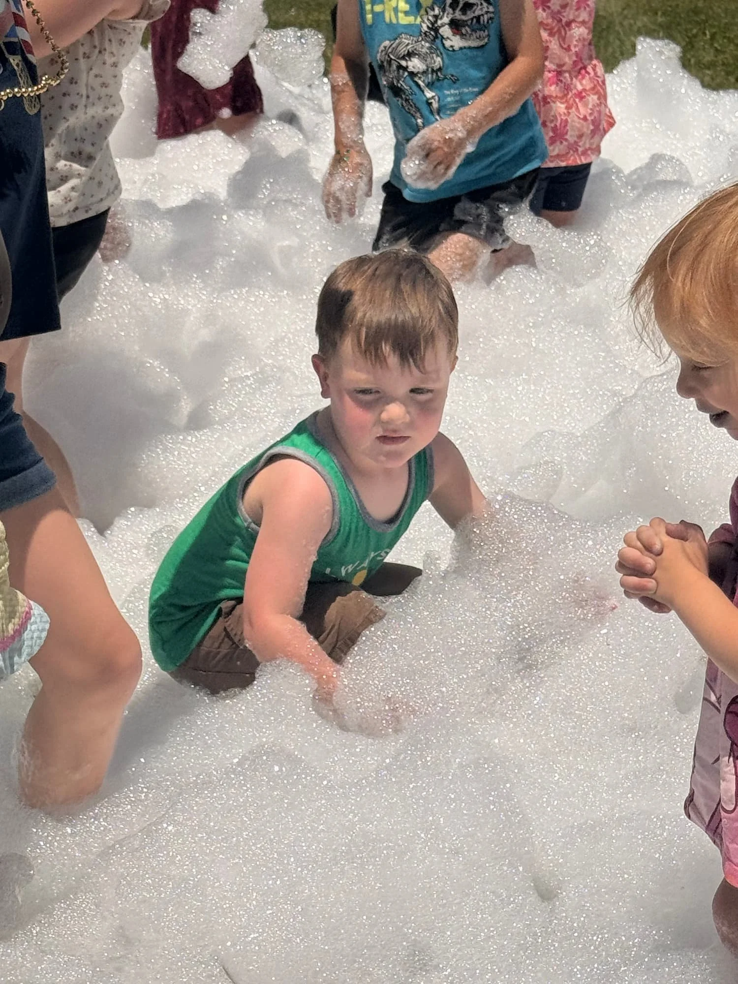 Children playing in a bubble-filled area, with one child sitting and grimacing at the camera while others stand or kneel around. The scene appears to be outdoors.