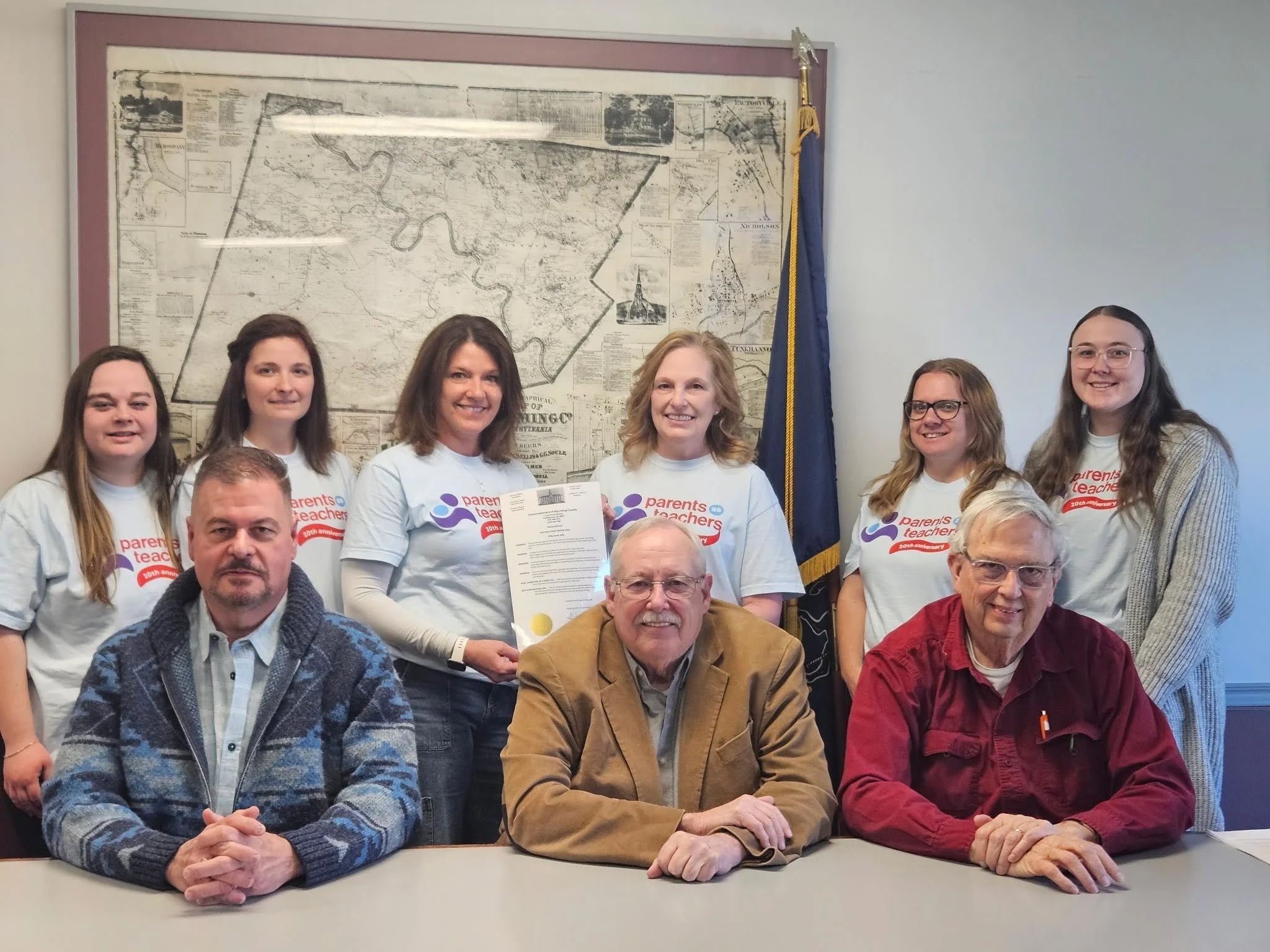 Group of people at a meeting, some holding certificates, in a room with a large map and a flag in the background.