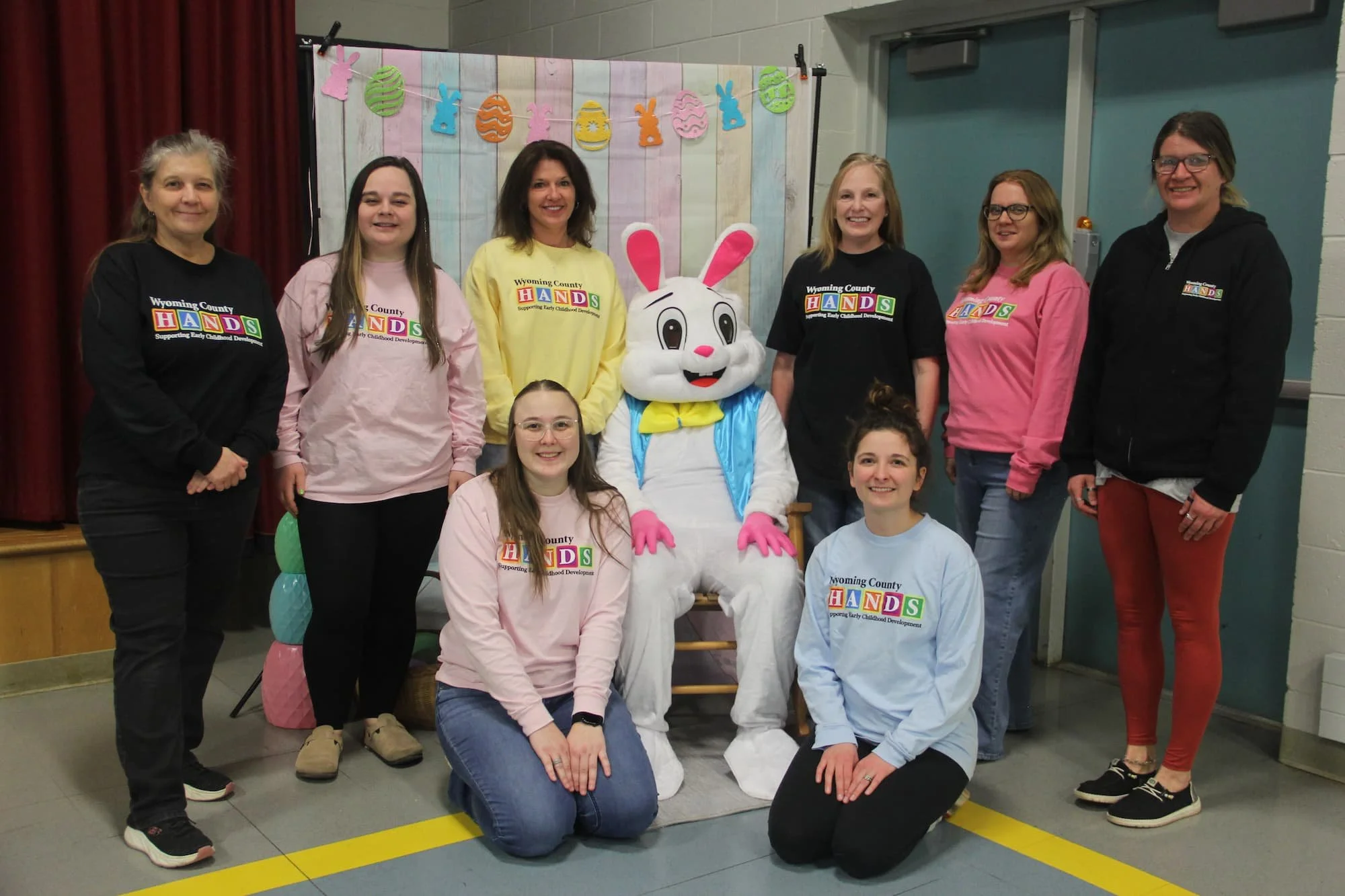A group of eight women and one person in a bunny costume are posing together indoors in front of a festive backdrop with colorful Easter-themed decorations. Some women are wearing Wyoming County H.A.N.D.S shirts.
