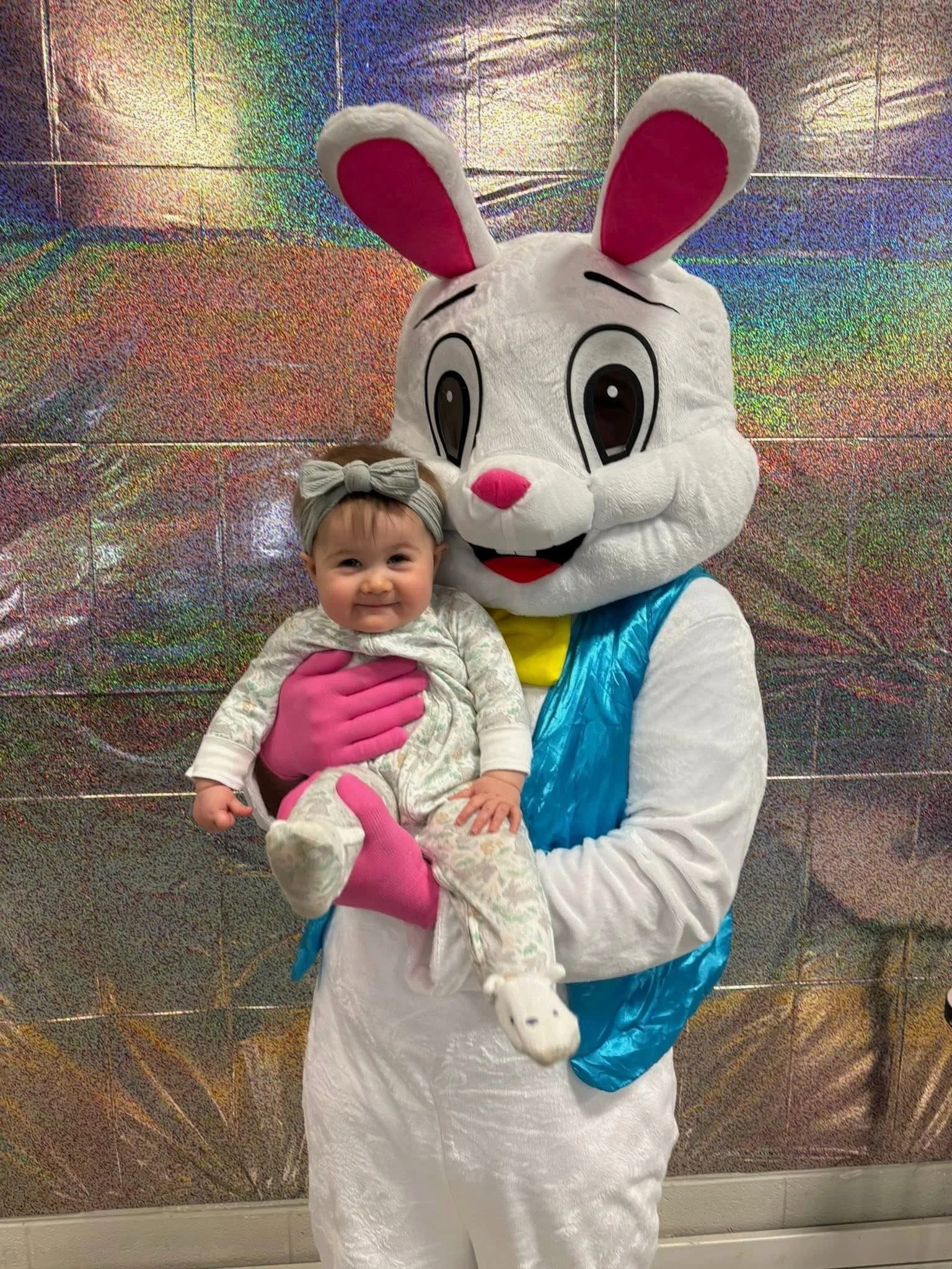 A young girl in pajamas sitting on the lap of a person in a large bunny costume. The costume bunny has a white furry body, big eyes, pink nose, and pink inner ears. The girl is wearing a gray headband with a bow and has a happy expression. The backgr