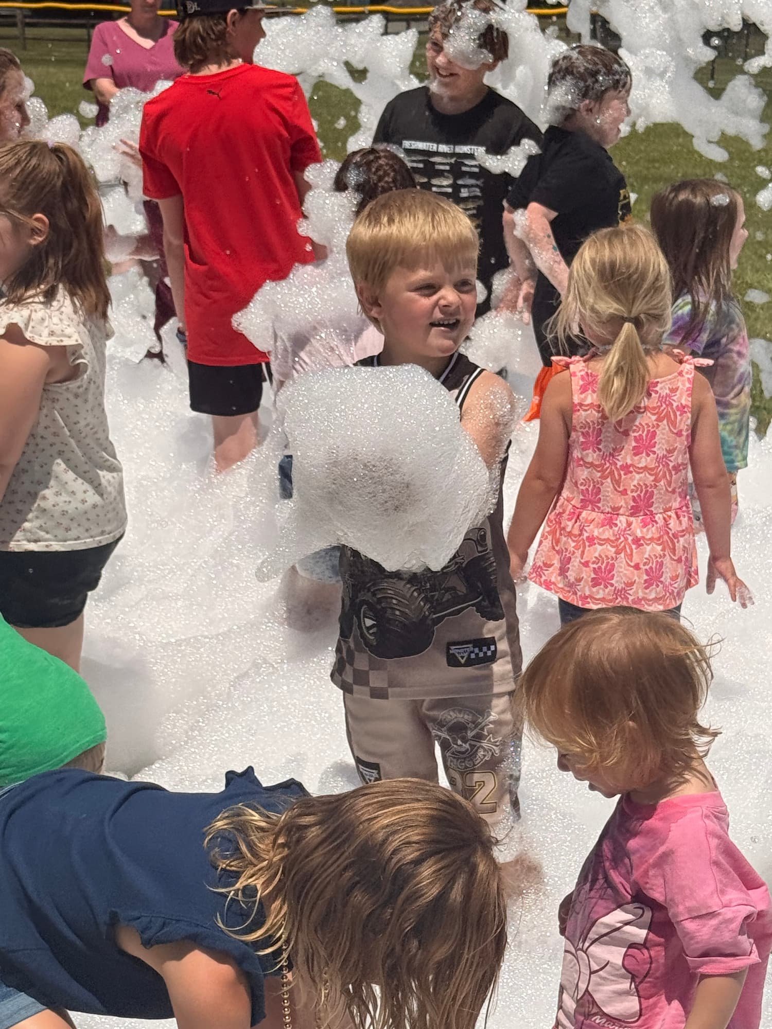 Children and adults playing with foam or bubbles outdoors on a grassy field, smiling and interacting with foam.