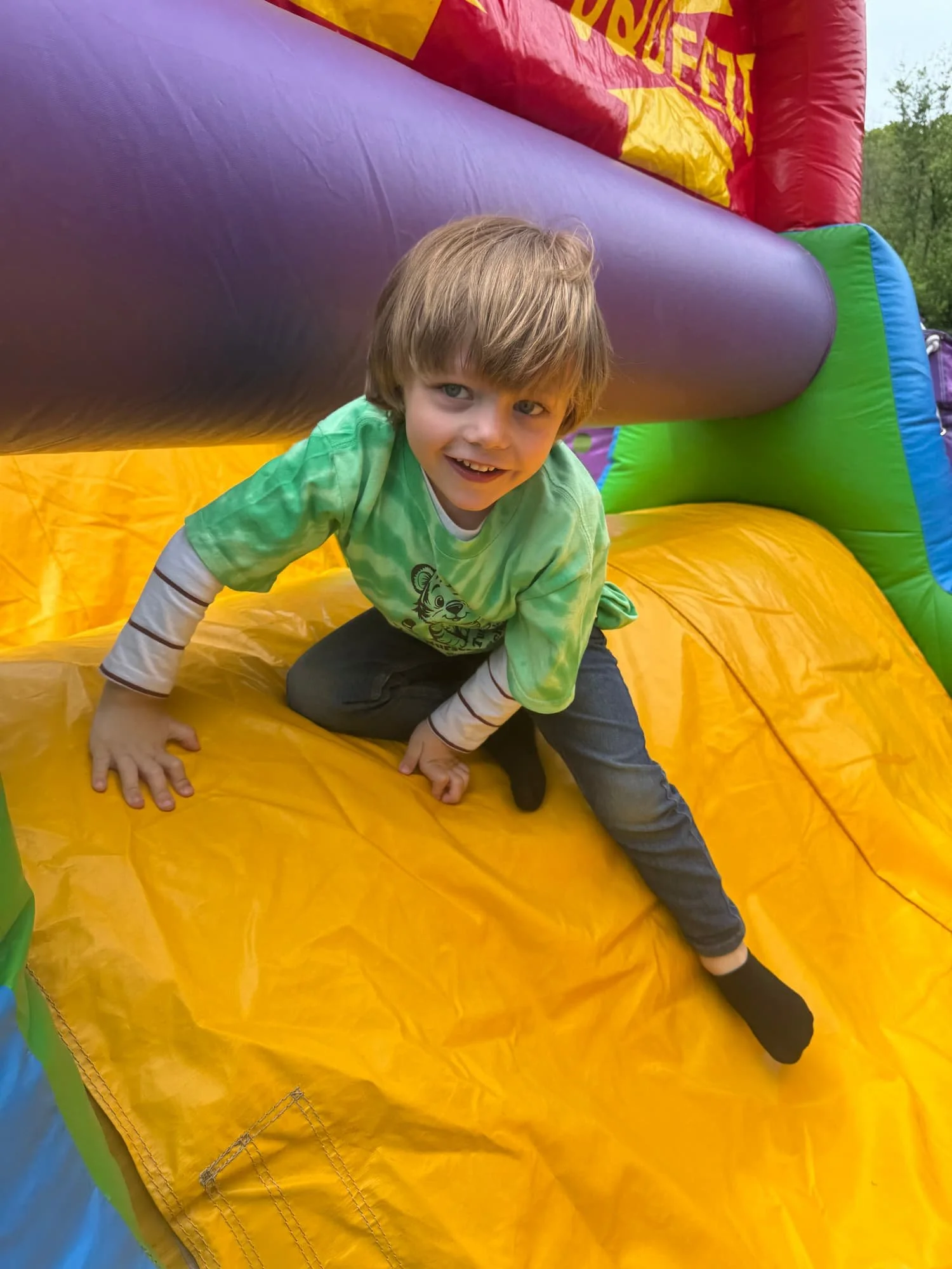 A young boy with brown hair and blue eyes crawling through a colorful inflatable slide at an outdoor event or park.