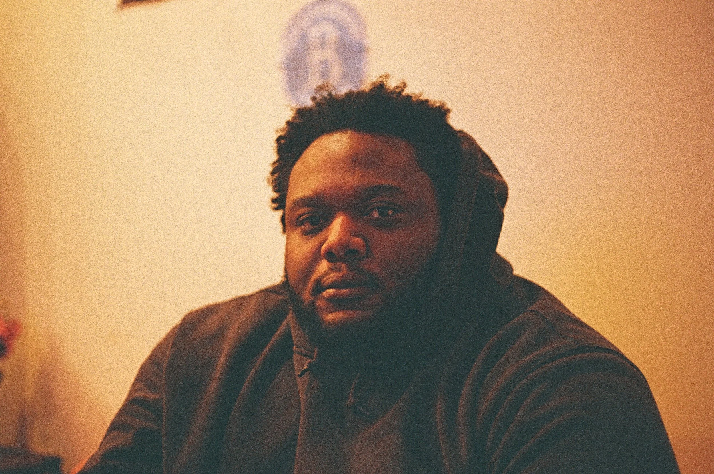 A young man with dark curly hair and facial hair, wearing a dark hoodie, sitting indoors with a neutral expression. The background is plain with a soft warm light.
