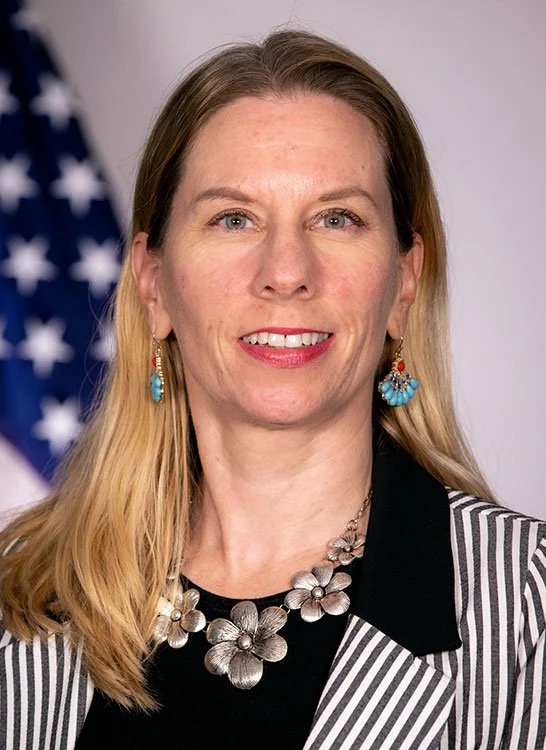 A woman with blonde hair and blue eyes, wearing patterned earrings and a floral necklace, standing in front of a blurred American flag.
