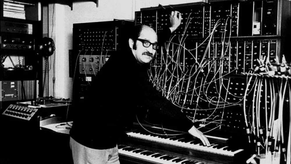 A man with glasses and a mustache working on an analog synthesizer, with numerous patch cables connected to the synthesizer modules in a studio or music production space.