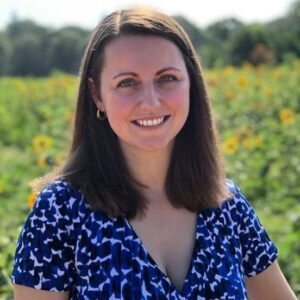A woman smiling outdoors in front of a field of sunflowers, wearing a blue patterned top and earrings.