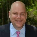A smiling man with a shaved head wearing a suit and pink tie, outdoors with green foliage in the background.