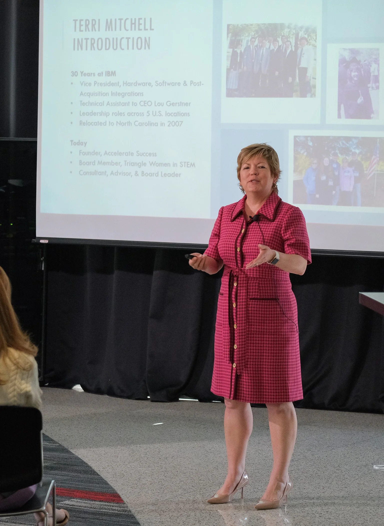 A woman with short blonde hair wearing a pink checkered dress and high heels presents in front of a projection screen. The screen displays her name, Terri Mitchell, and her professional background and achievements. There are photos on the screen of people, possibly at previous events.