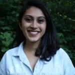 A young woman with dark hair smiling outdoors, wearing a light-colored shirt, against a background of green trees.