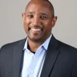 A smiling African American man in a dark suit jacket and light blue shirt, posing against a plain background.