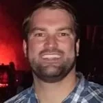A smiling man with short brown hair and a beard, wearing a button-up shirt, in a dimly lit environment with red lighting in the background.