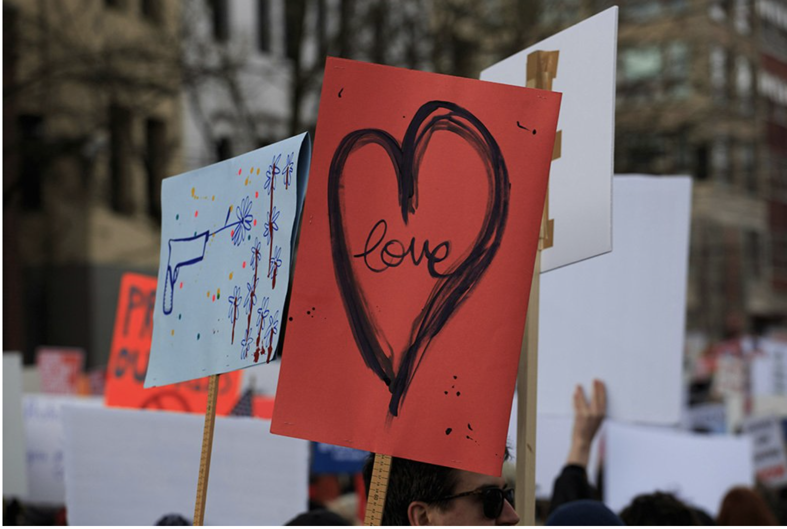 Multiple signs and posters held up by a crowd of people at an outdoor event. Visible signs include one with a red background with a heart outline and the word 'love' written inside, and another with a light blue background featuring doodles of flowers and abstract designs.
