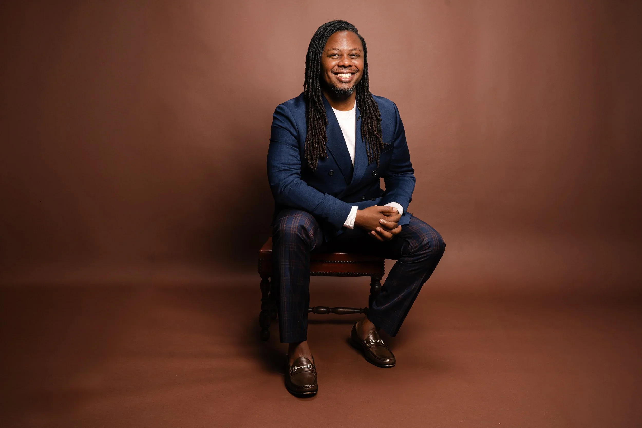 A man with long dreadlocked hair and a beard, smiling while sitting on a wooden chair against a plain brown background. He is wearing a double-breasted blue blazer, plaid pants, and brown loafers.