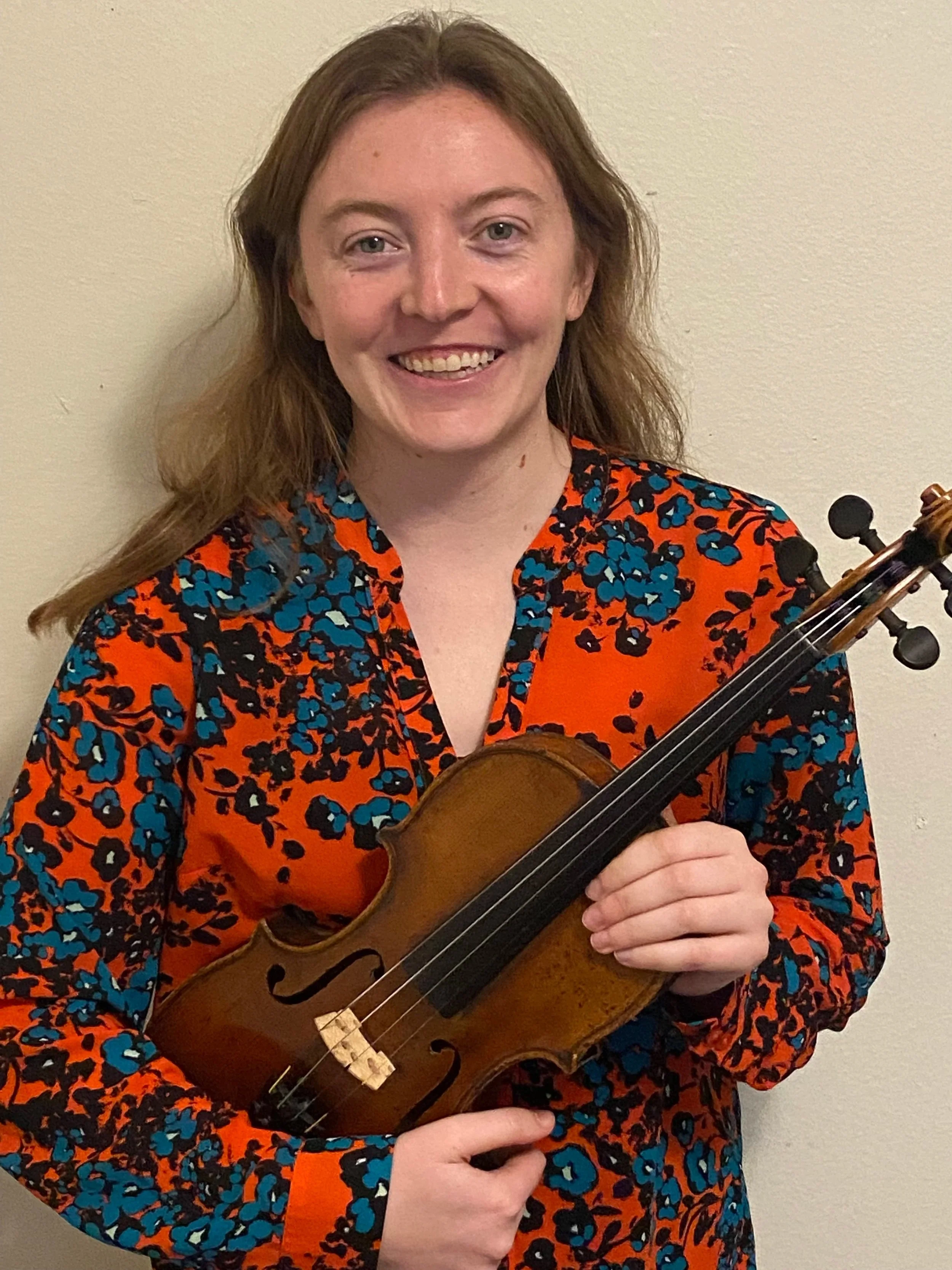 A woman with long brown hair smiling and holding a violin, wearing a vibrant orange and blue patterned shirt, standing against a plain light-colored wall.
