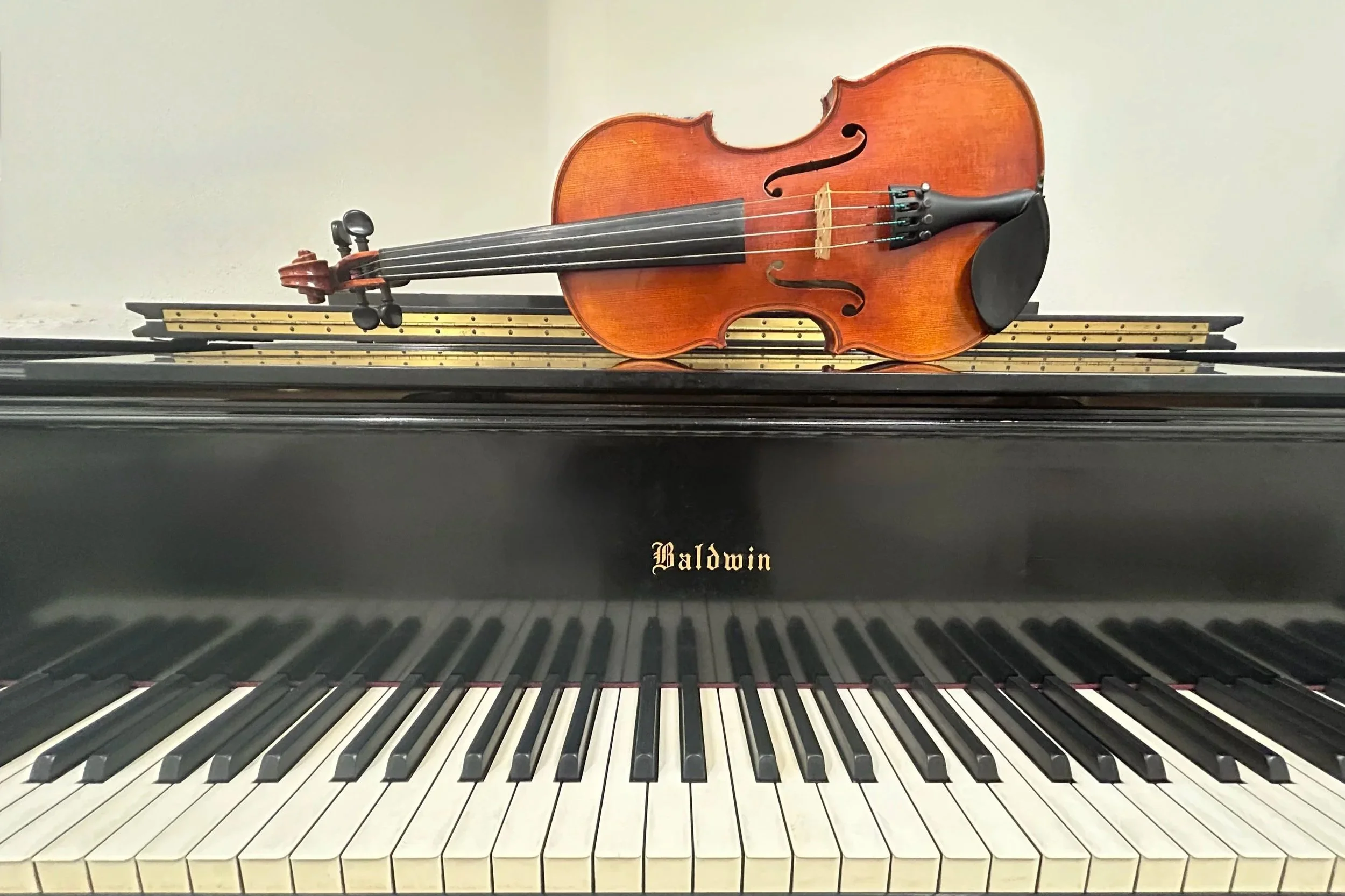 A close-up view of a violin resting on a black piano with white and black keys. The violin is placed horizontally on top of the piano. The brand name 'Baldwin' is visible on the piano.