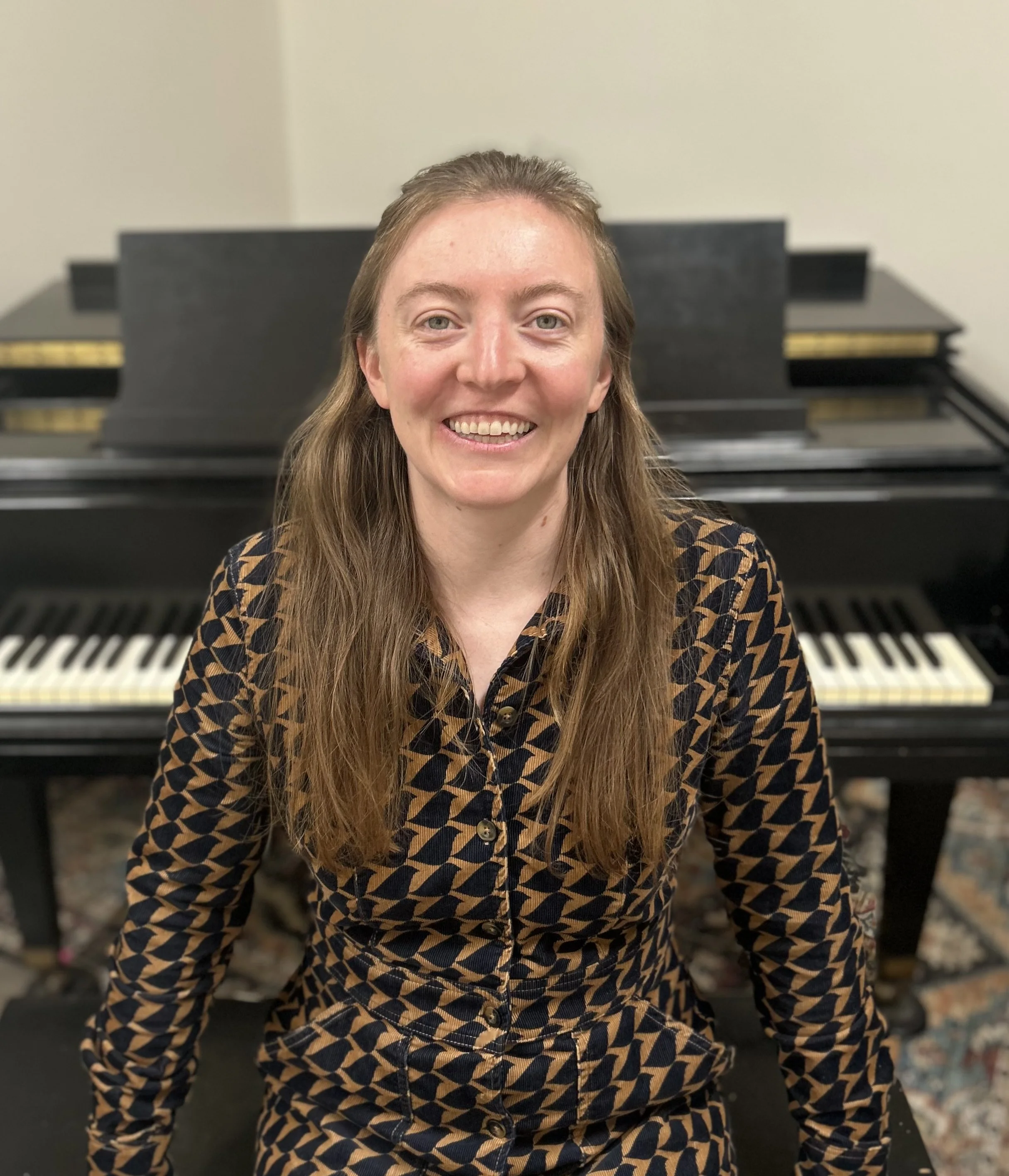A smiling woman with long, light brown hair, wearing a patterned black and gold shirt, sits in front of a piano.