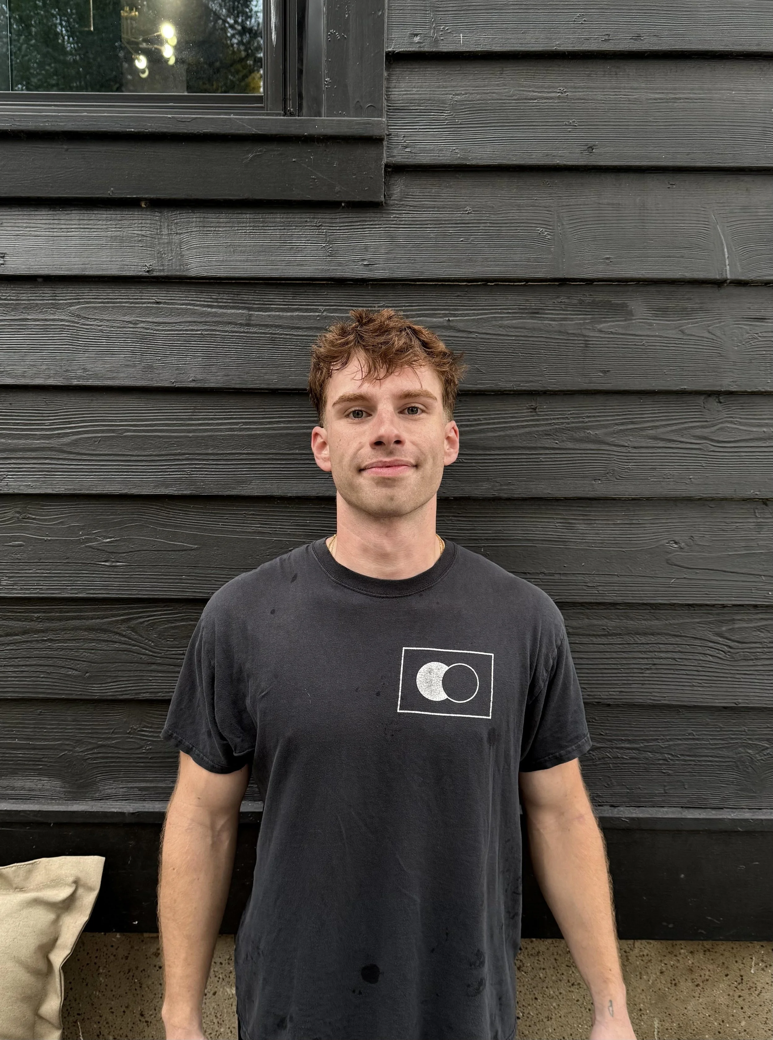 A young man with short, curly brown hair and light skin, wearing a black t-shirt with a white geometric design, stands in front of a dark wooden wall. Part of a beige bag is visible on the left side of the image.