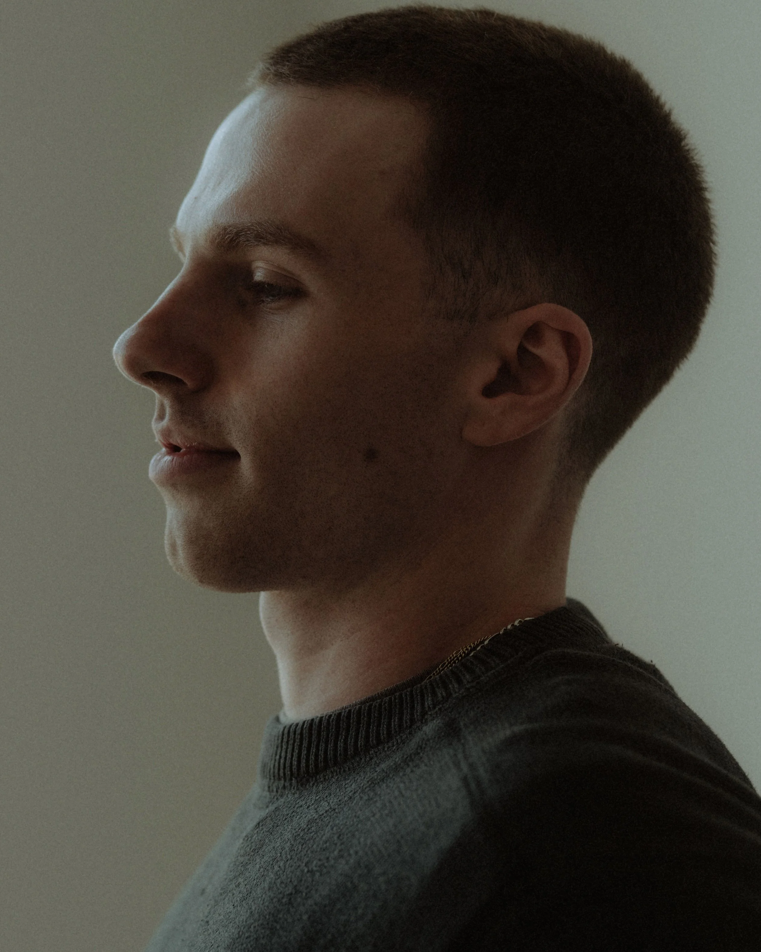 Side profile of a young man with short brown hair, wearing a dark-colored top with a ribbed collar.