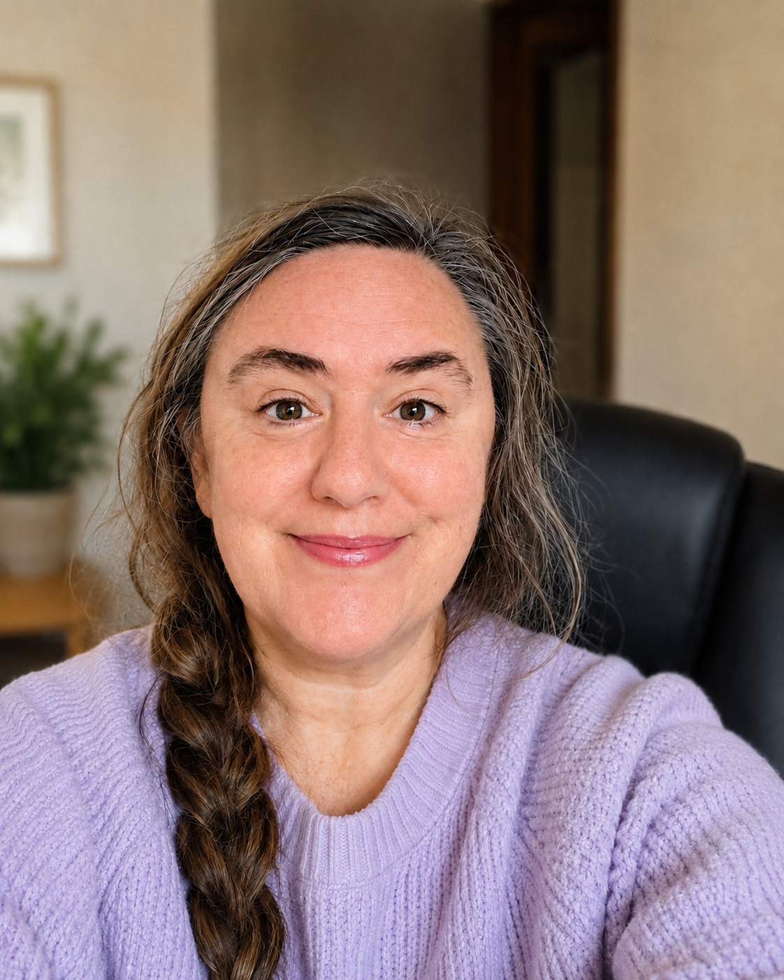 Close-up of a smiling woman with long wavy brown hair, wearing a light purple sweater, sitting in an office chair in a warmly lit room.