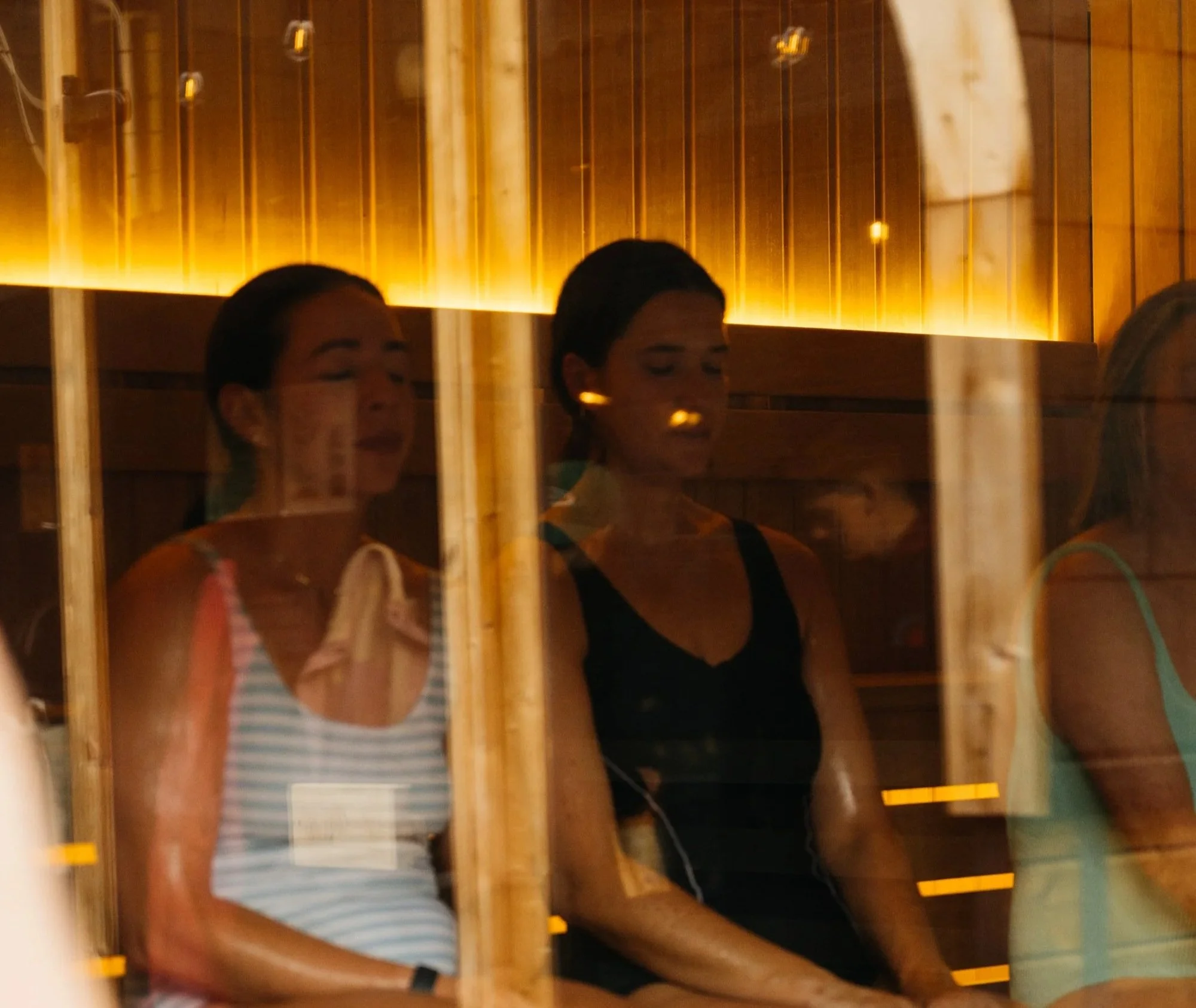 Two women are taking a selfie in a mirror at a gym. They are both dressed in workout clothes, with one in a striped tank top and the other in a black top. The gym has wooden walls with yellow lighting, and there are yellow markers on the mirror edges.