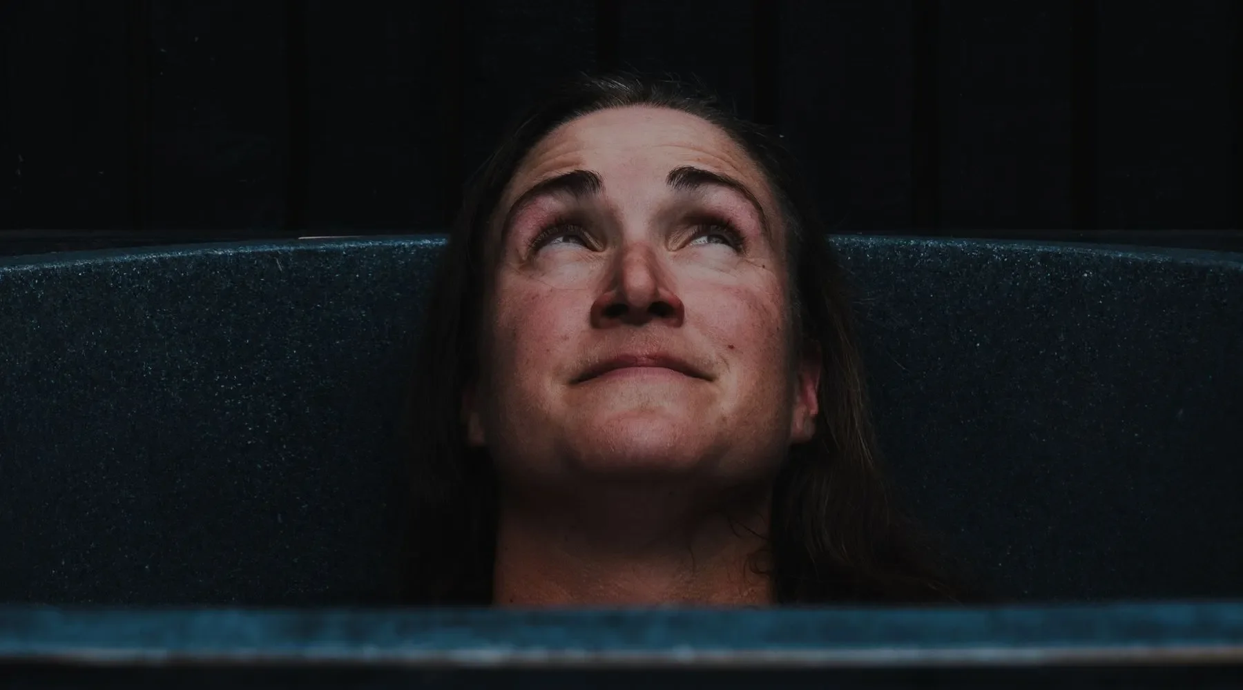 A woman with dark hair relaxing in a water tub, looking up with a peaceful expression.