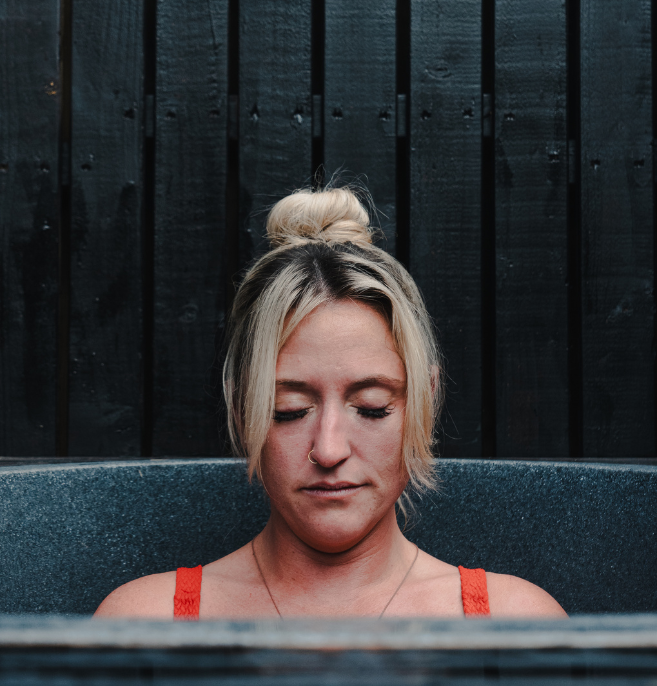 Woman with blonde hair in a bun, eyes closed, sitting in a hot tub with a dark wooden fence in the background.