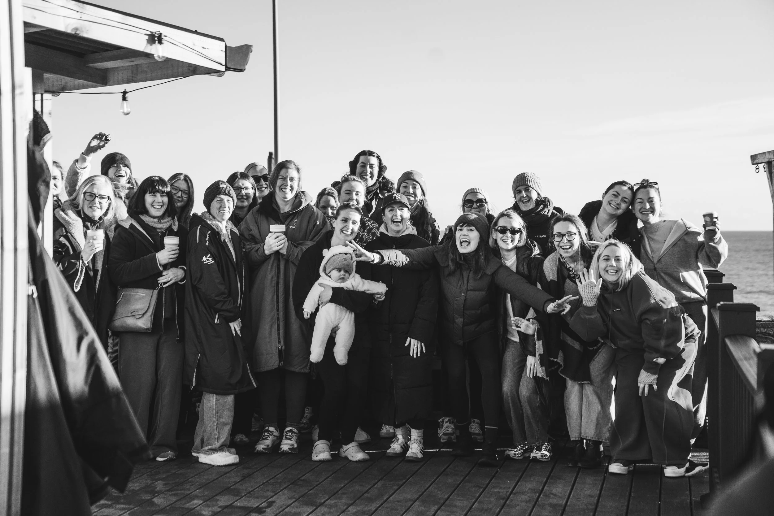 A group of people smiling and laughing on a boat deck, holding drinks, with some wearing winter clothing like hats and jackets.