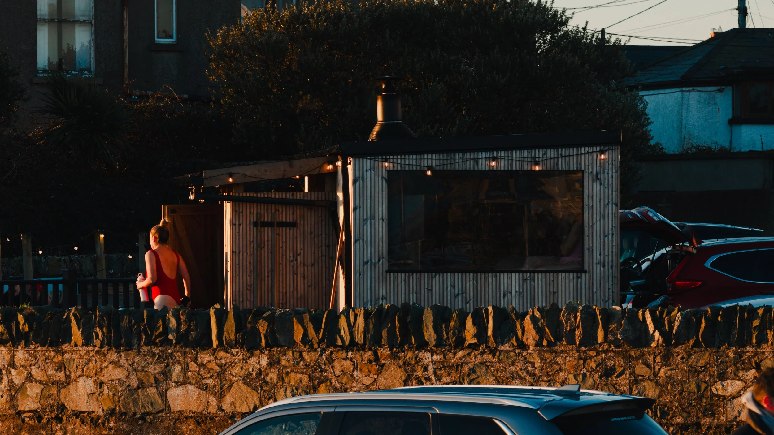 A woman in a red dress holding a drink stands outside a small, rustic building decorated with string lights, during dusk.