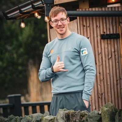 A young man with glasses wearing a light blue long sleeve shirt standing outdoors near a wooden fence, making a shaka sign with his right hand and smiling.