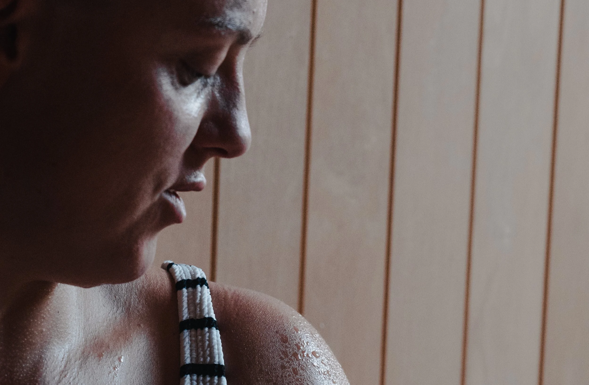 Close-up of a person's face and shoulder from the side, with sweat on their skin, wearing a striped tank top, against a wooden wall backdrop.