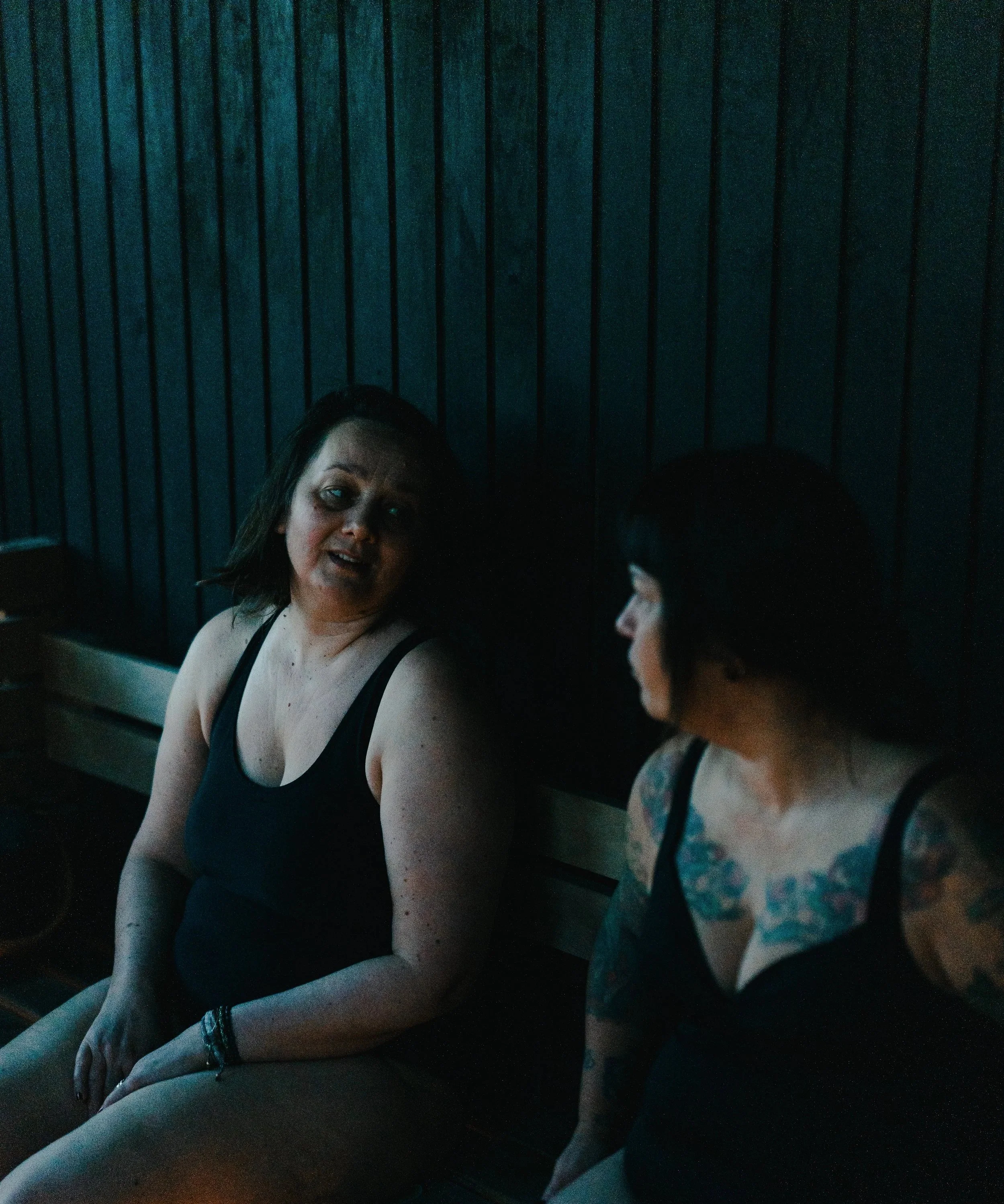 A woman with dark hair, wearing a black tank top, sitting at a bar or restaurant, engaged in conversation with woman in floral top. The background has vertical dark wood paneling.