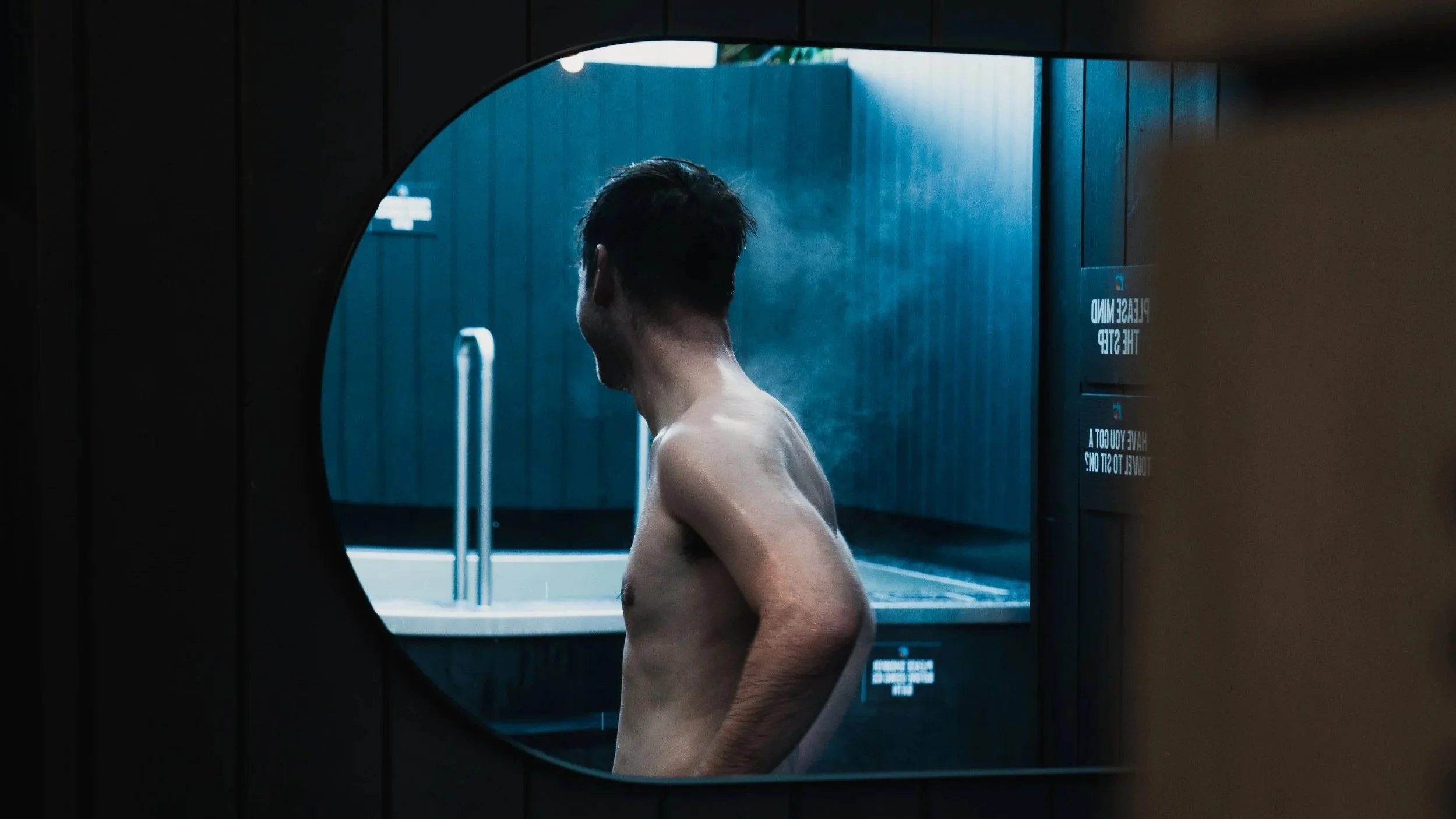 A shirtless man with wet hair standing in front of a blue wall, seen through a circular window or mirror.