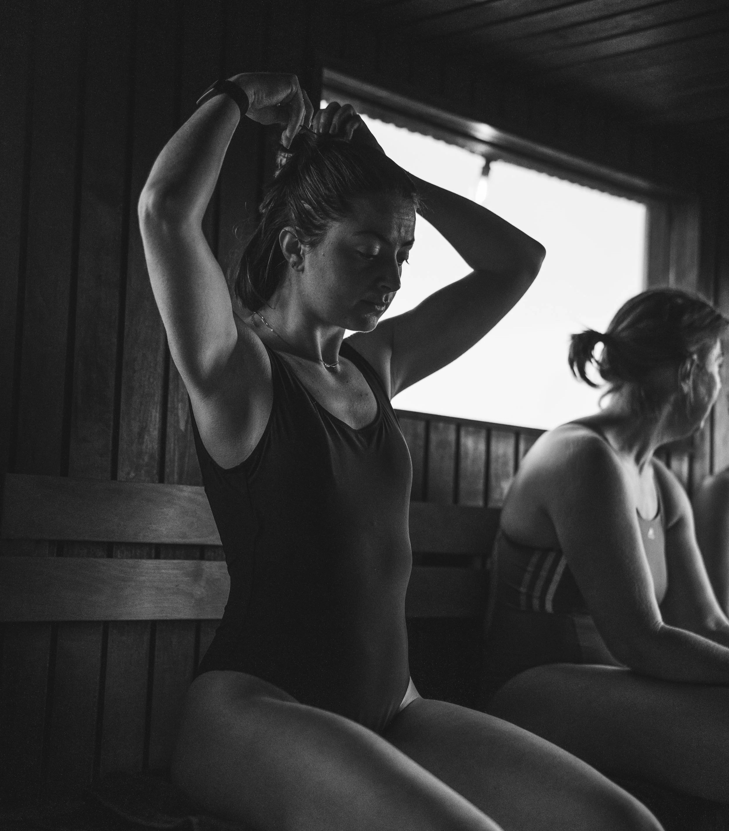 Black and white photo of a woman in workout clothes stretching her hair with her hands, sitting near a window, with wooden wall panels in the background.
