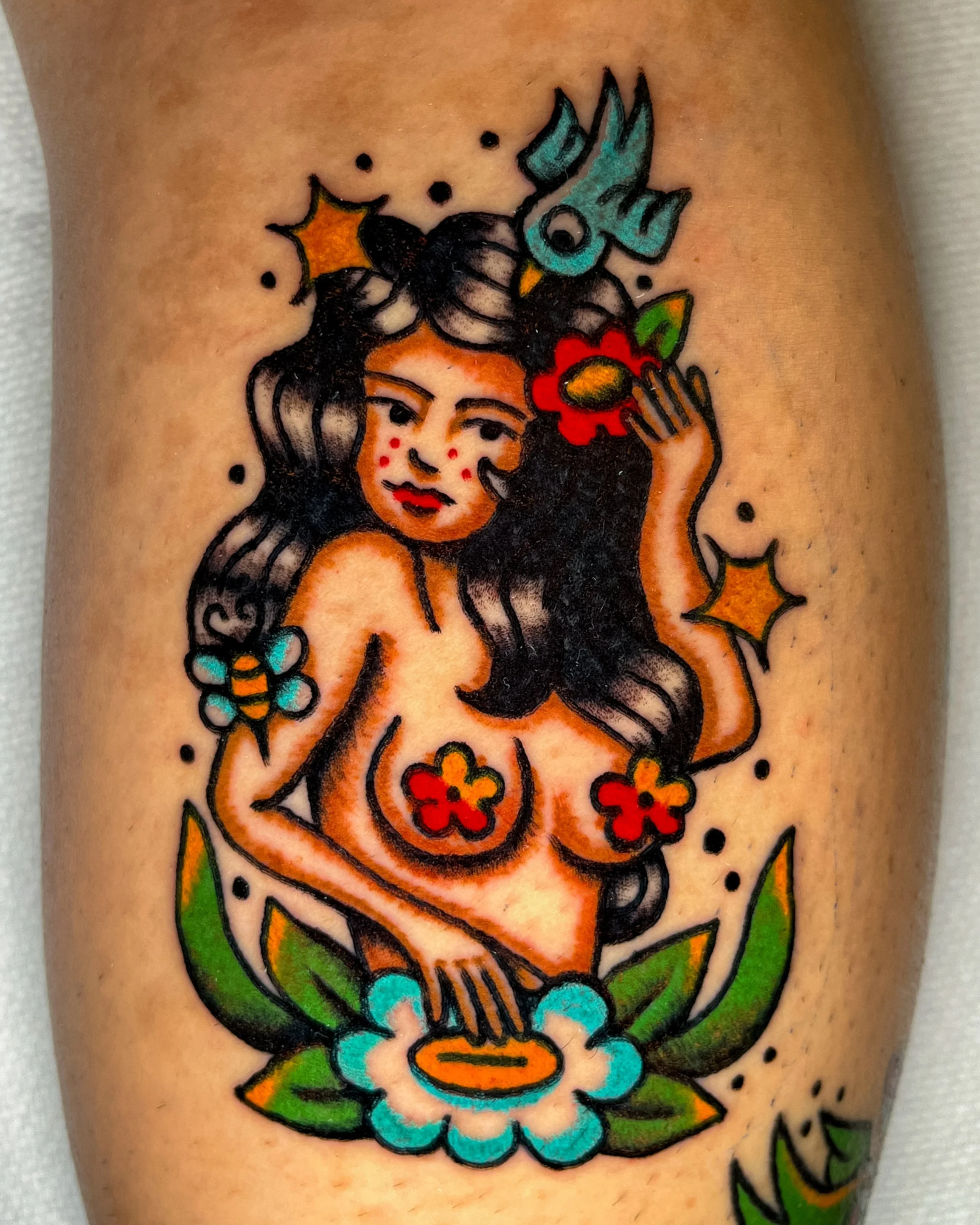 Colorful tattoo of a woman with long dark hair, freckles, and orange lipstick, holding a red flower near her face, surrounded by green leaves, yellow stars, and blue butterflies.