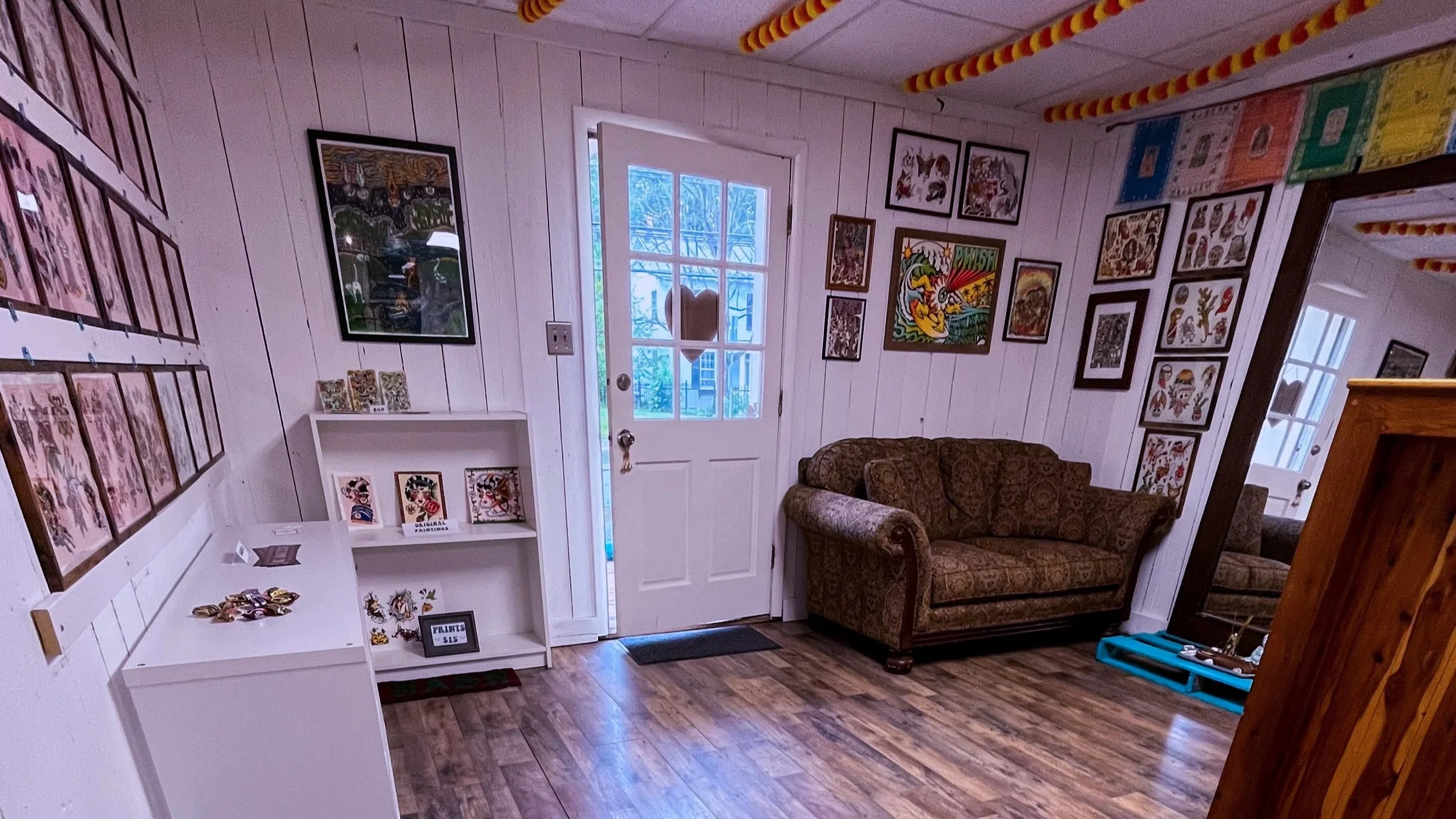 A cozy indoor space with white painted wooden paneled walls decorated with various framed artwork. There is a white shelf displaying small items and a sign that reads "PRINTS $15." A brown patterned couch sits against the wall, and a door with glass panes leads outside, allowing natural light into the room. The wooden floor adds warmth to the setting.