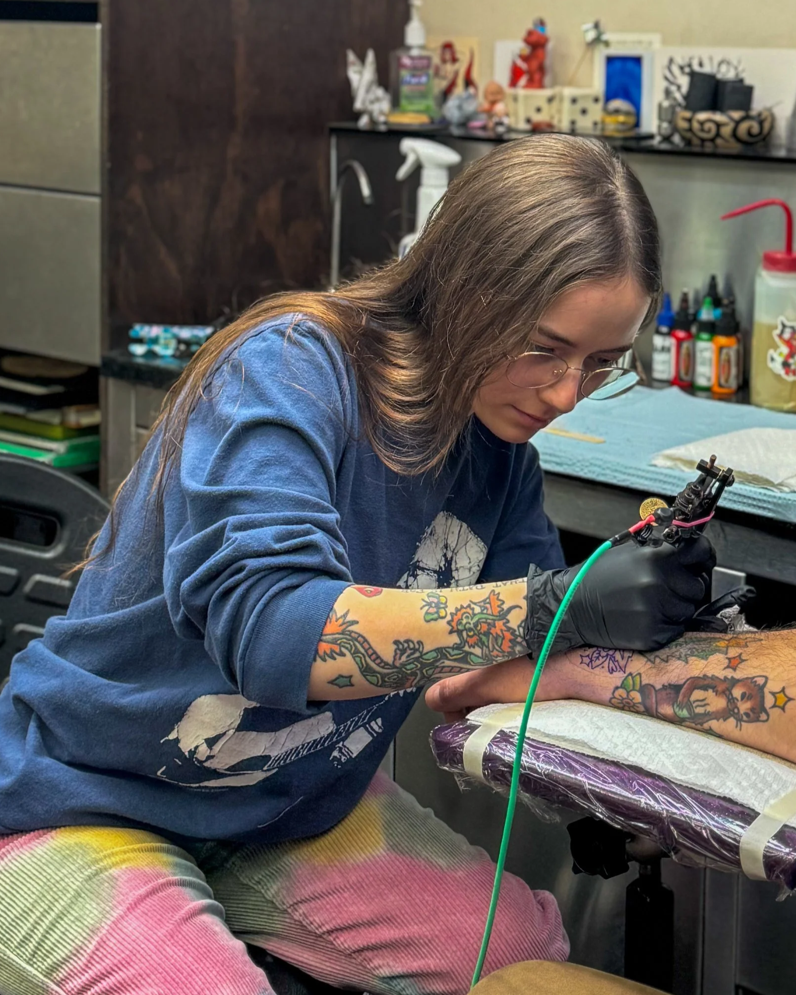A person with long hair and glasses working on a tattoo machine, wearing a colorful tattoo sleeve on their arm in a tattoo studio.