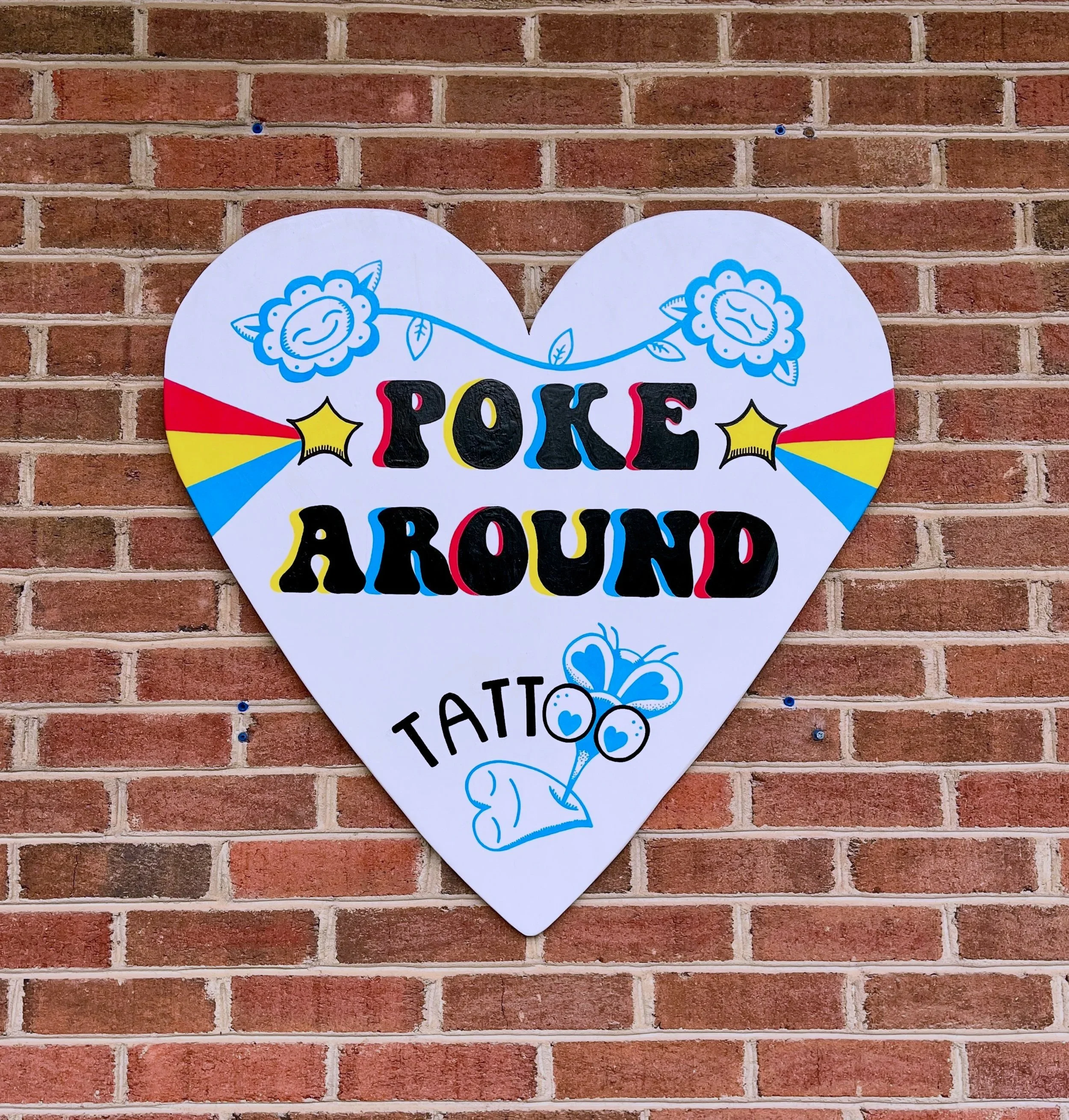 A heart-shaped decorative sign with smiling cartoon flowers, colorful text that reads 'POKE AROUND,' and blue and yellow accents, mounted on a brick wall.