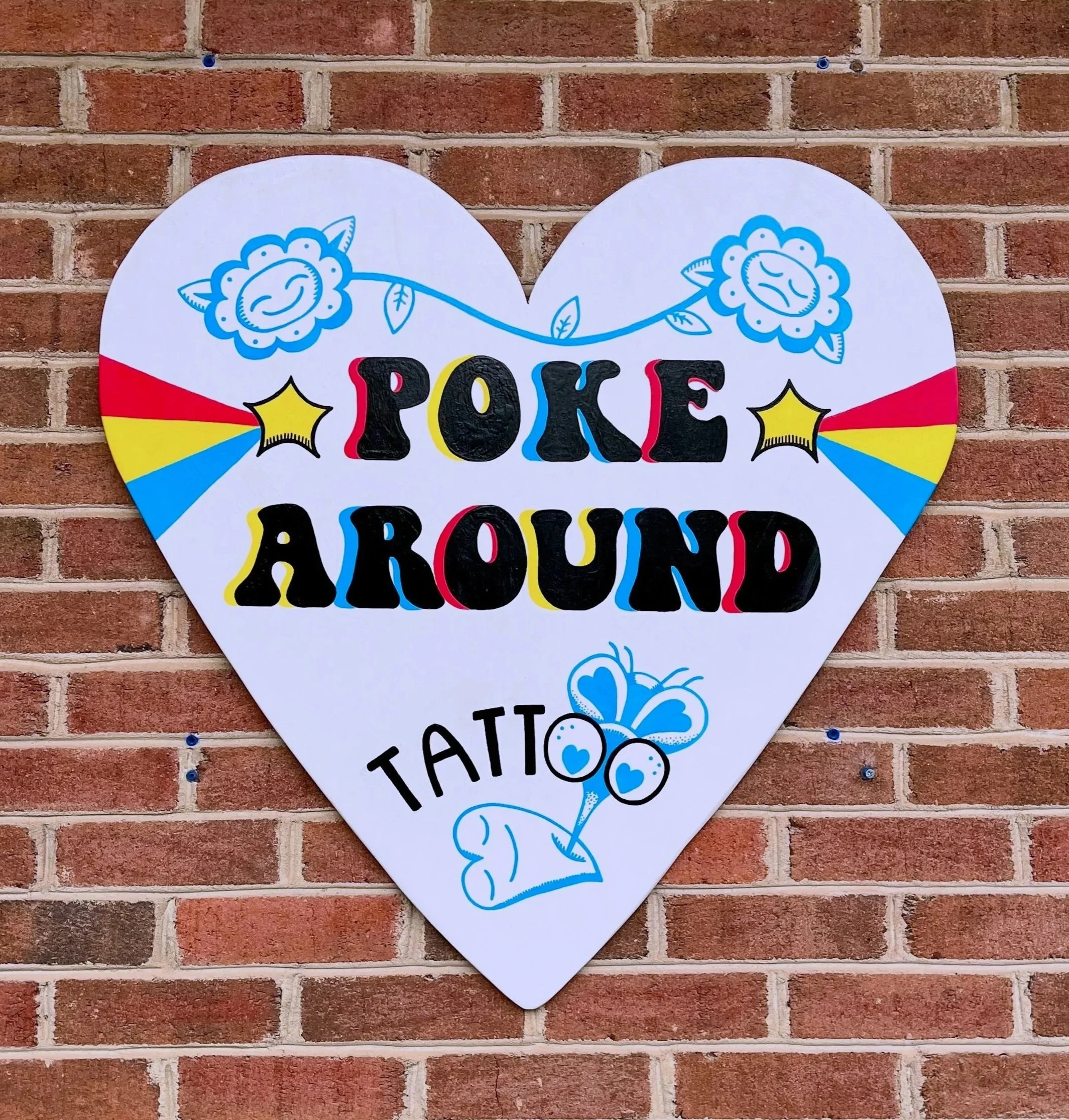 Heart-shaped sign with colorful text and illustrations, including blue flowers and a bee, attached to a brick wall.