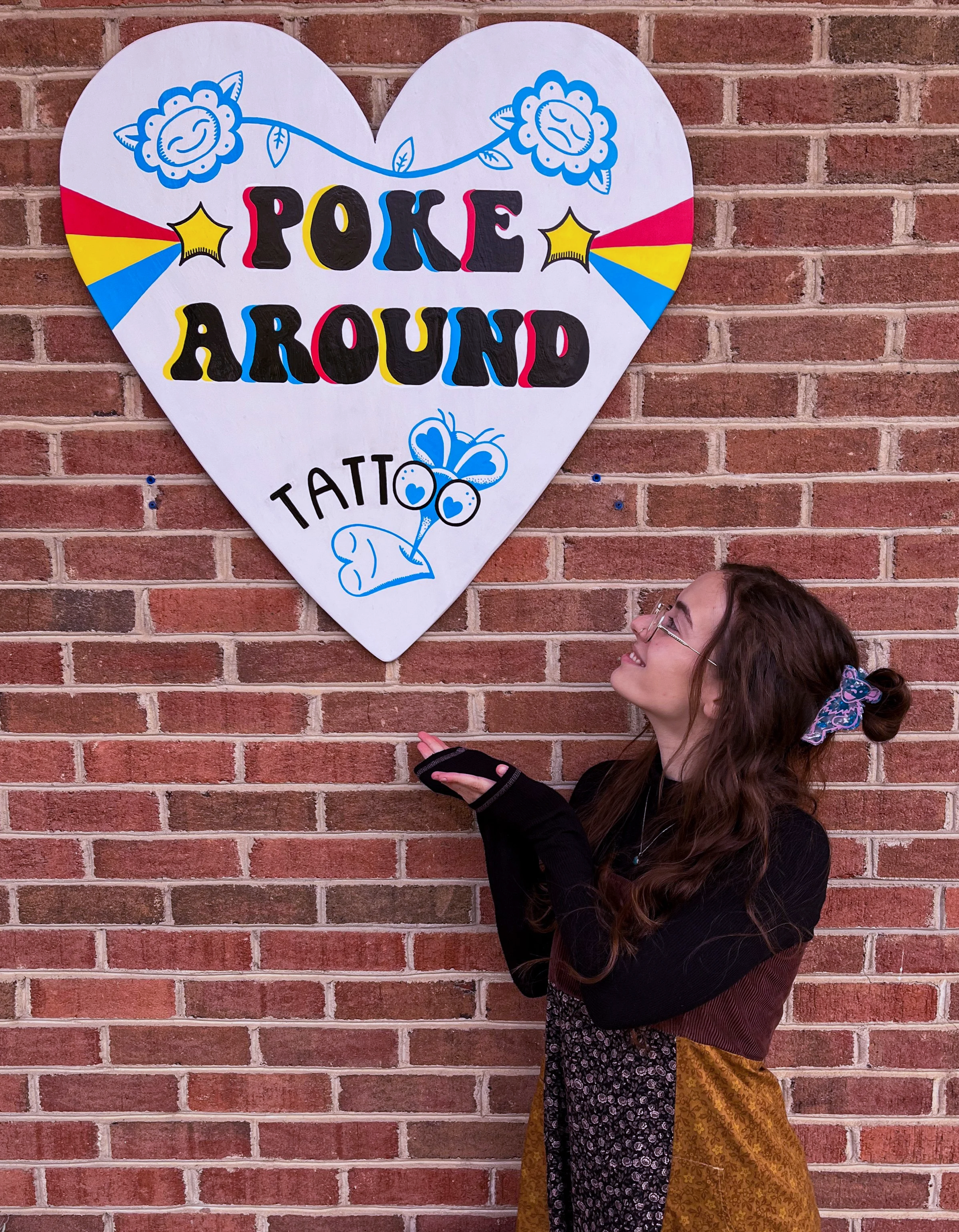 A woman with glasses and long hair tied with a floral scrunchie, smiling and pointing at a heart-shaped sign on a brick wall. The sign has the words 'POKE AROUND' in bold colorful letters, along with drawings of blue flowers, stars, a butterfly, and bright color accents.
