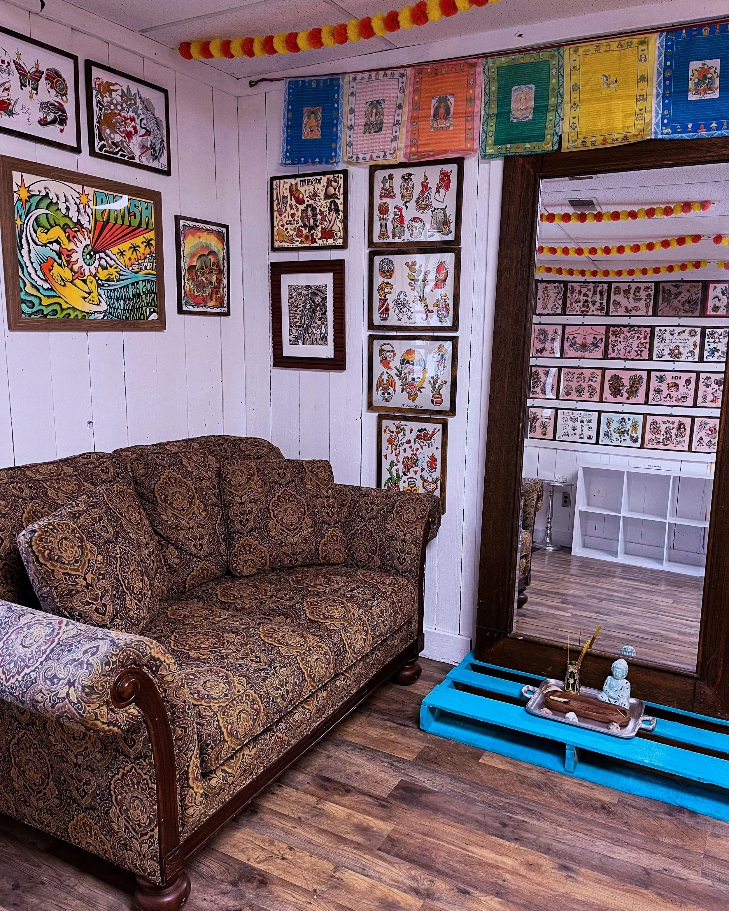 A cozy room decorated with colorful artwork on white paneled walls and a row of multicolored paper garlands hanging from the ceiling. The room features a vintage-patterned sofa with ornate wood trim, situated next to a large mirror framed in dark wood. The reflections in the mirror reveal more artwork and part of another room. On the floor, a bright blue pallet holds a tray with small decorative statues and items, next to a small white shelving unit.