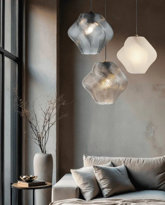 Modern living room with three hanging pendant lights, a beige sofa with multiple pillows, a small round side table with a plant and books, next to a large window with natural light, and a tall white vase with dry branches nearby.