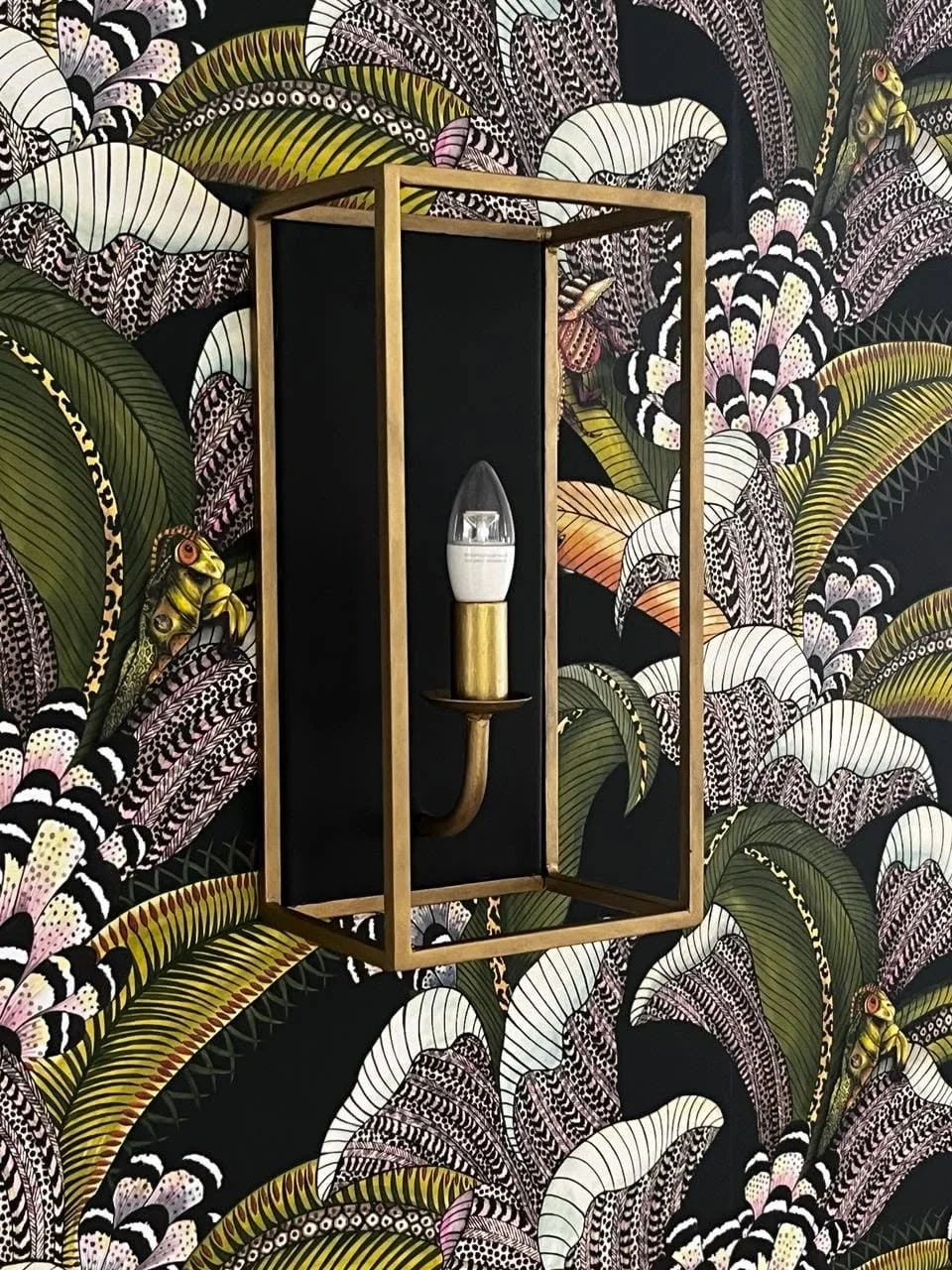 A modern wall sconce with a black backplate, gold frame, and white bulb with a clear cover, mounted on a wallpapered wall with a tropical leaf and animal print pattern.
