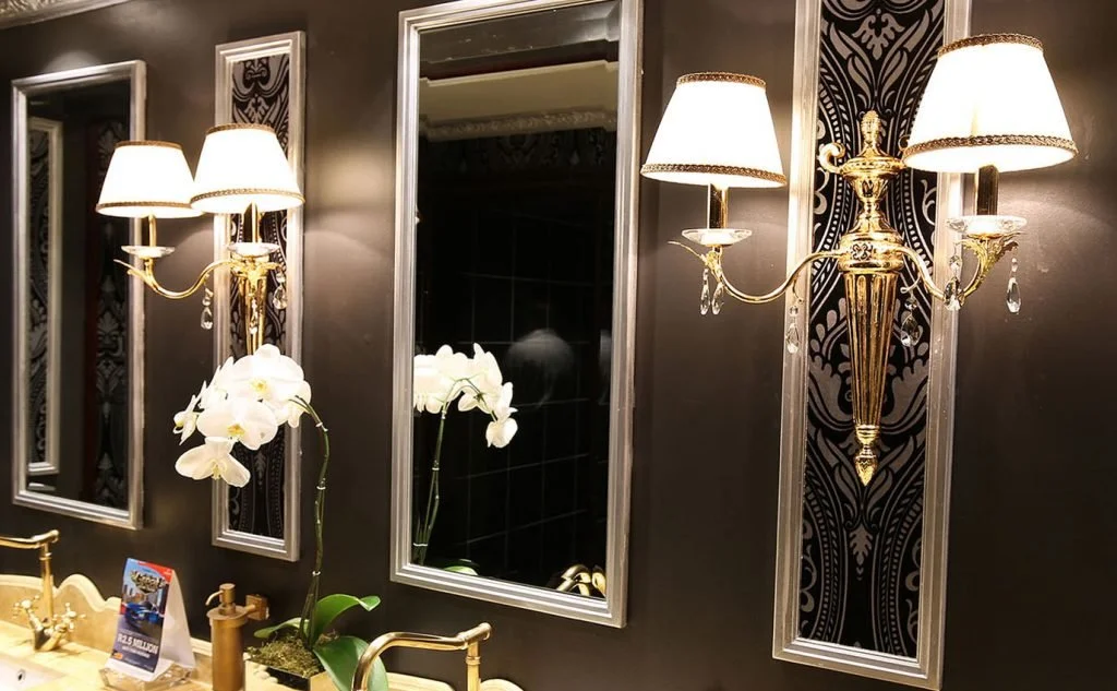 Elegant bathroom interior with three large framed mirrors, gold wall sconces with lampshades and hanging crystal accents, and a vase with white orchids on a gold sink counter.