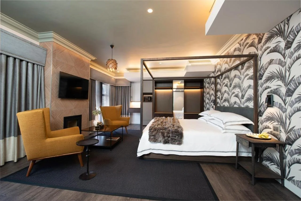 Modern hotel room with a four-poster bed, patterned wallpaper, yellow armchairs, a small side table with a plant, a wall-mounted TV, and large windows with curtains.